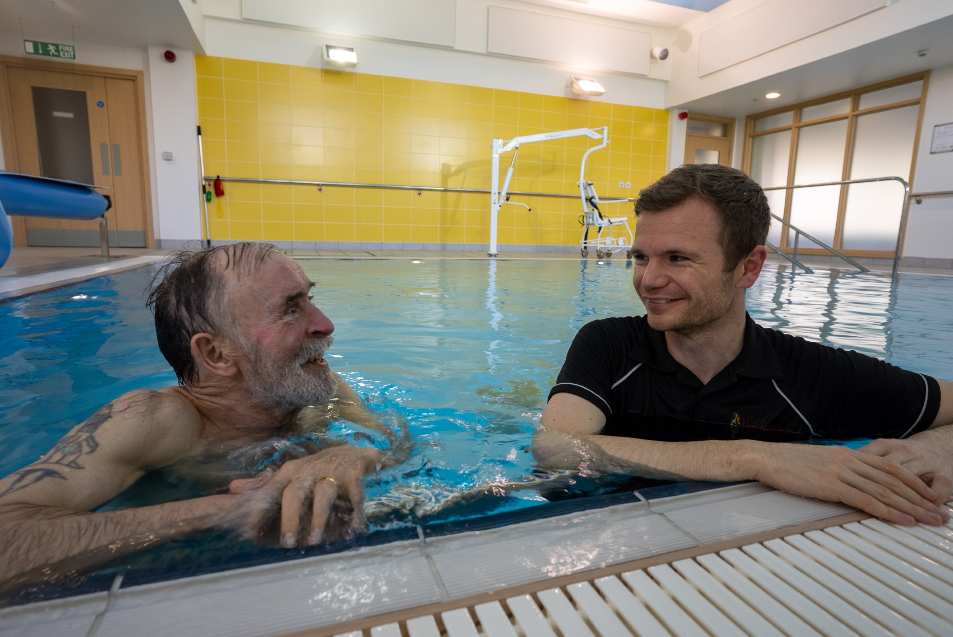 An older man and a younger man talk and smile while standing in an indoor swimming pool. The room has yellow tiles and a pool lift is visible in the background.