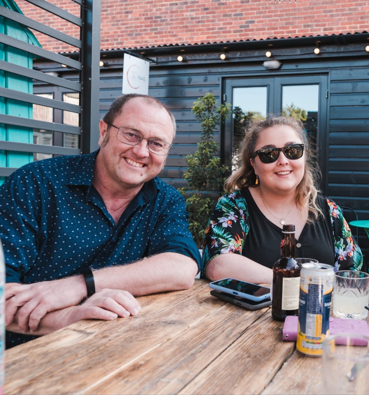 Two people sit at an outdoor wooden table, smiling. The man wears glasses and a blue shirt; the woman has sunglasses and a floral top. Drinks and a phone are on the table. A black building and plants are in the background.