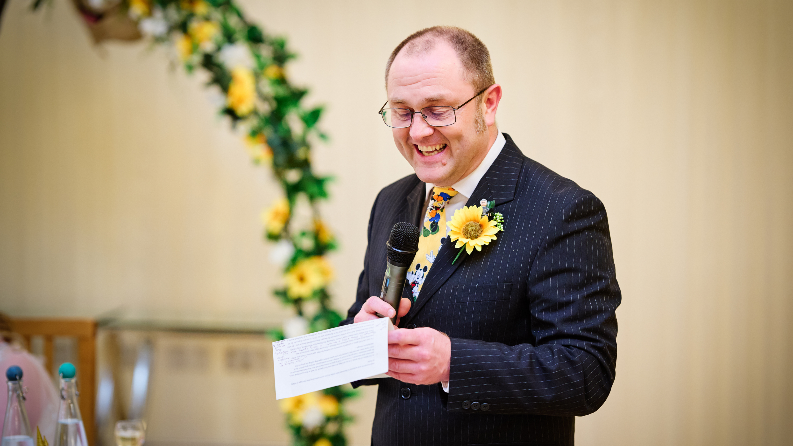 A man in a suit with a sunflower boutonnière smiles while speaking into a microphone and holding a paper, standing beside a floral arch decorated with yellow flowers.