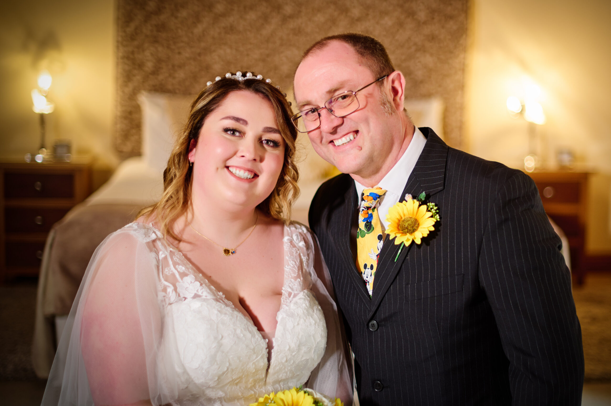 A bride in a white dress and a man in a suit with a sunflower boutonniere smile together indoors, with a bed and warm lighting in the background.