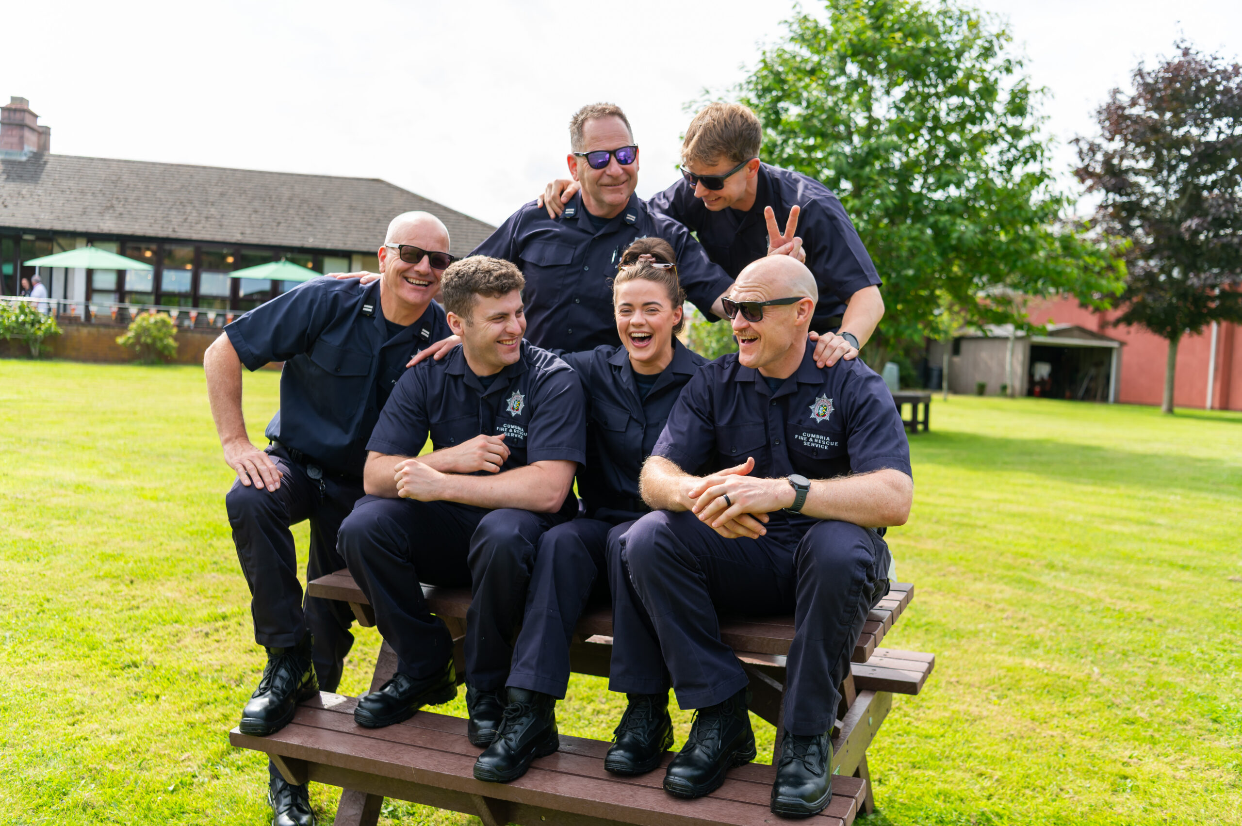 Five people in matching navy uniforms sit and laugh together on a wooden picnic table outdoors, with green grass and trees in the background. One person playfully makes bunny ears over another's head.