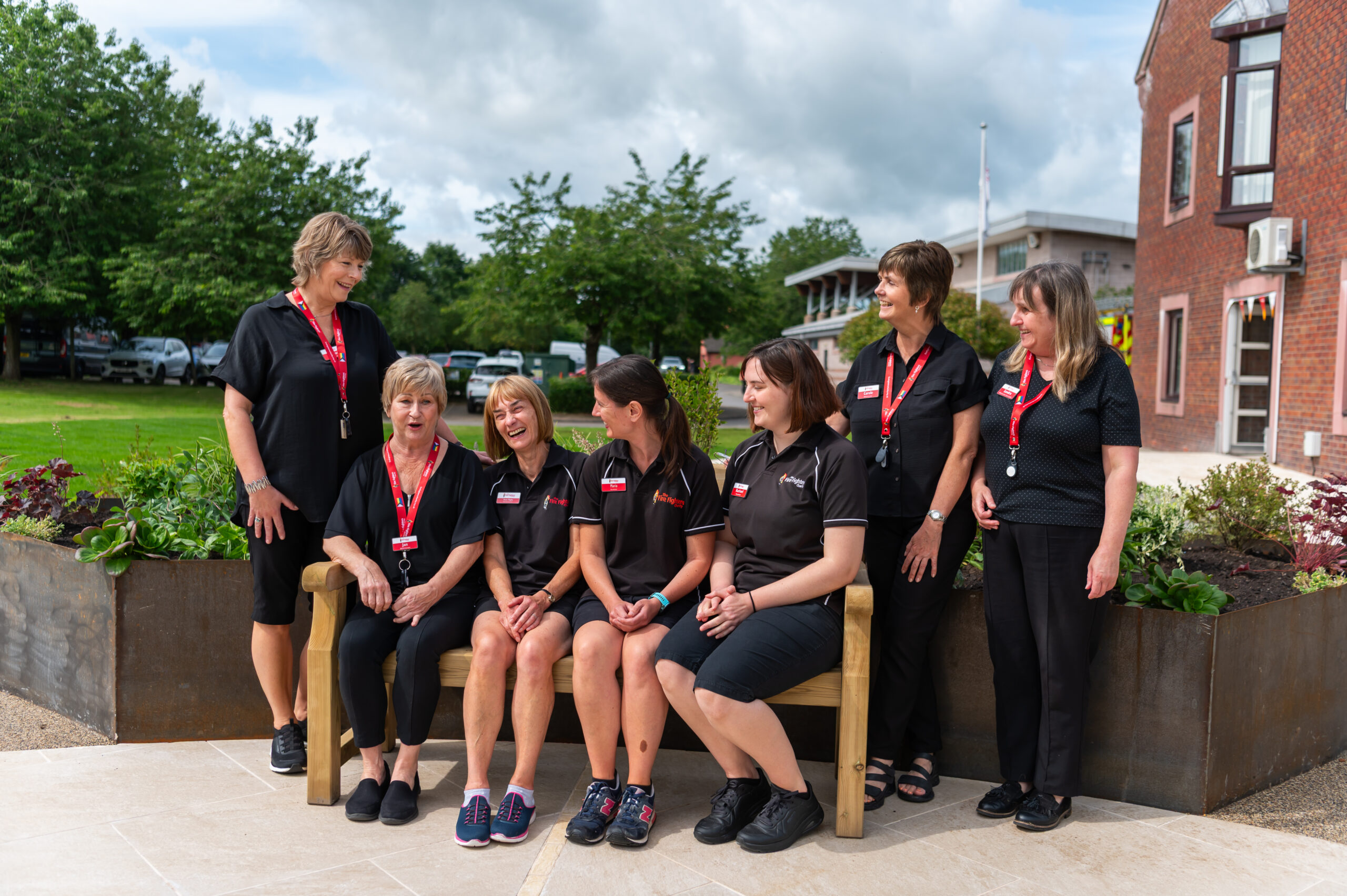 Seven women in matching black uniforms and red lanyards, representing a fire service charity, pose outside on a sunny day; six stand while one sits on a wooden bench, and they are smiling and laughing together in front of greenery and a brick building.