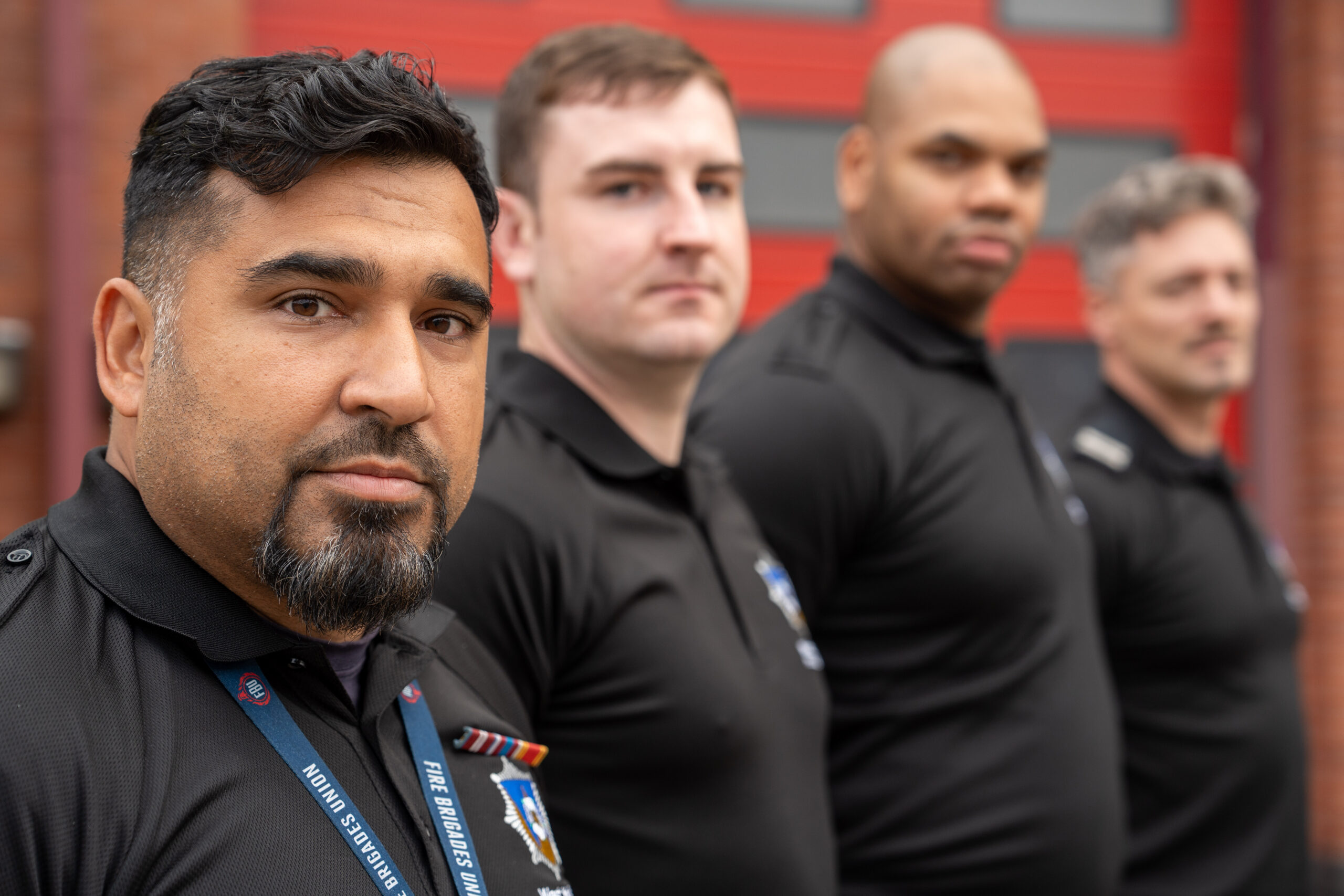 Four men in black uniforms stand in a row outdoors, facing the camera. The focus is on the man in front, while the others are slightly out of focus. A red door and brick wall are in the background.