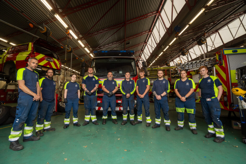 Ten firefighters in uniform stand in a row inside a fire station, with fire trucks and equipment visible in the background under a high, metal roof.
