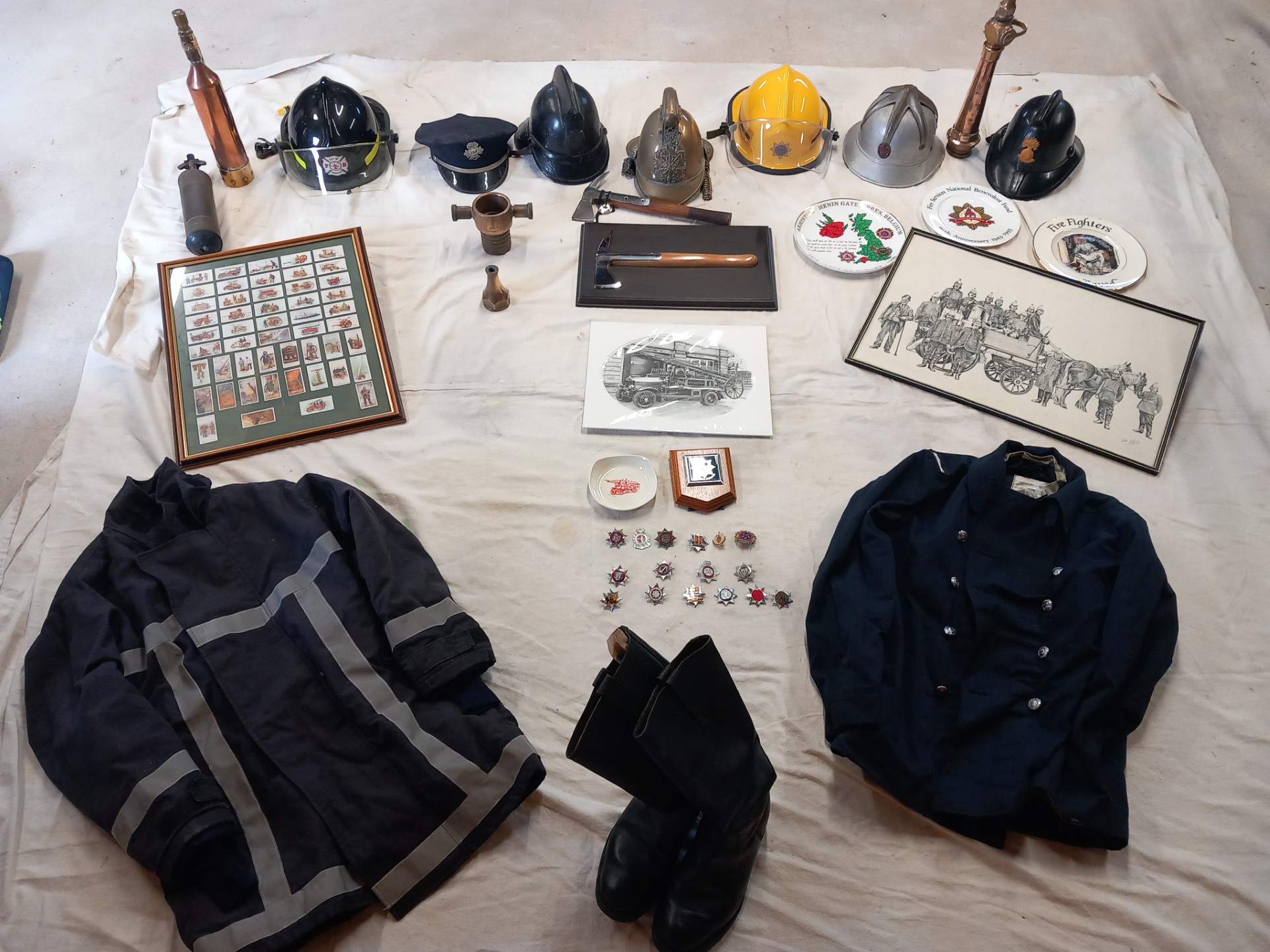 A collection of vintage firefighting gear, helmets, boots, jackets, awards, framed badges, ceremonial items, and illustrations neatly arranged on a white cloth.