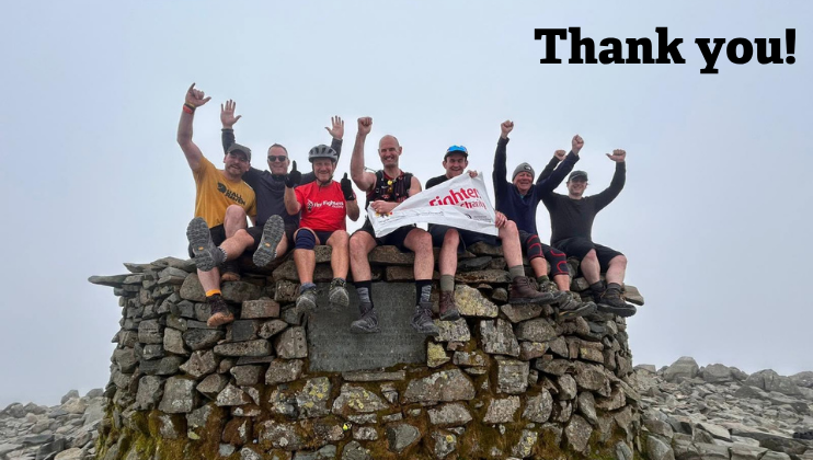A group of eight people sit on a stone structure outdoors, raising their arms in celebration. They hold a banner and smile. The sky is overcast, and 