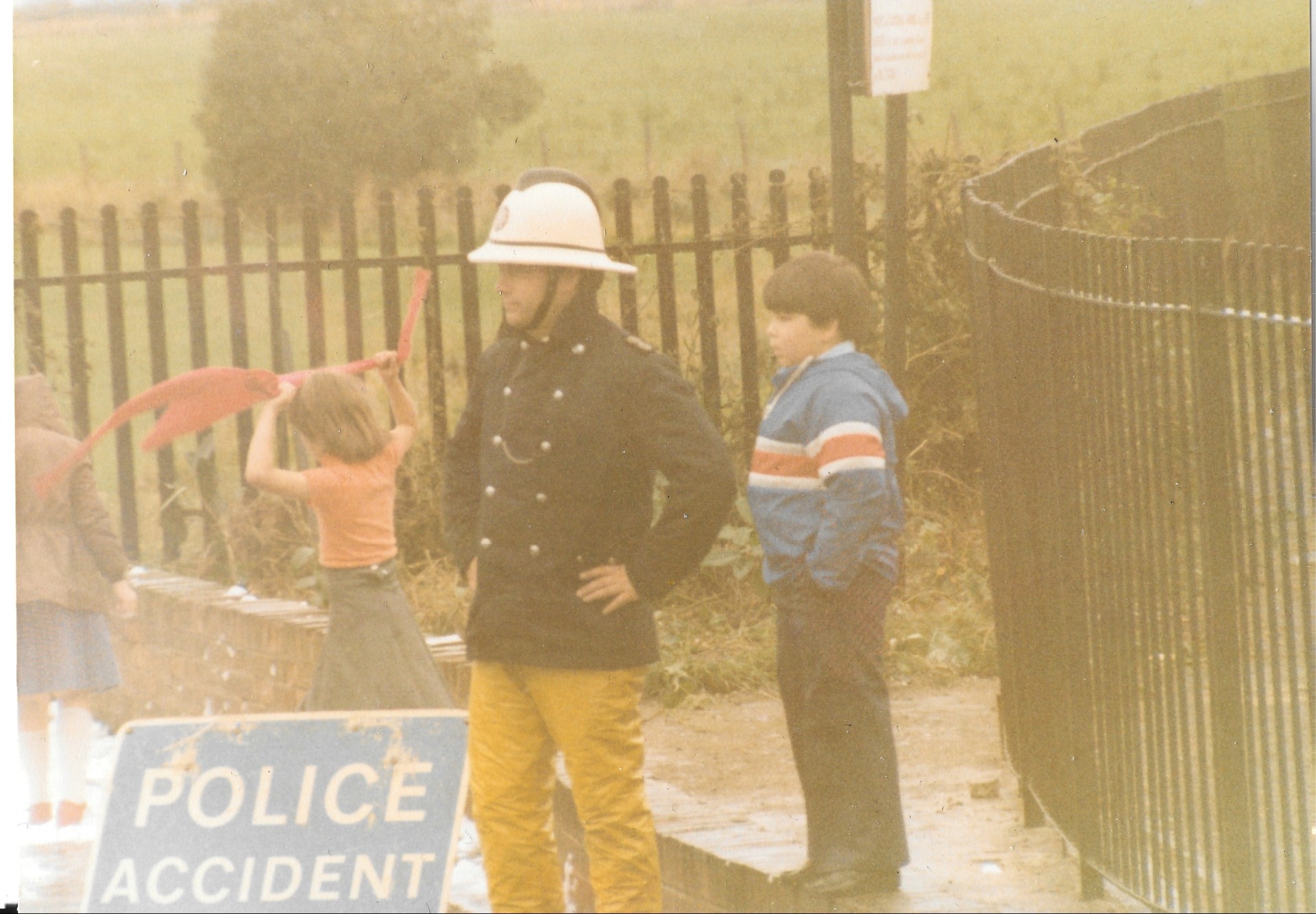 A firefighter in a helmet and yellow pants stands beside two children near a “Police Accident” sign and a black metal fence. One child waves red tape. The scene appears to be outdoors on a rainy day.