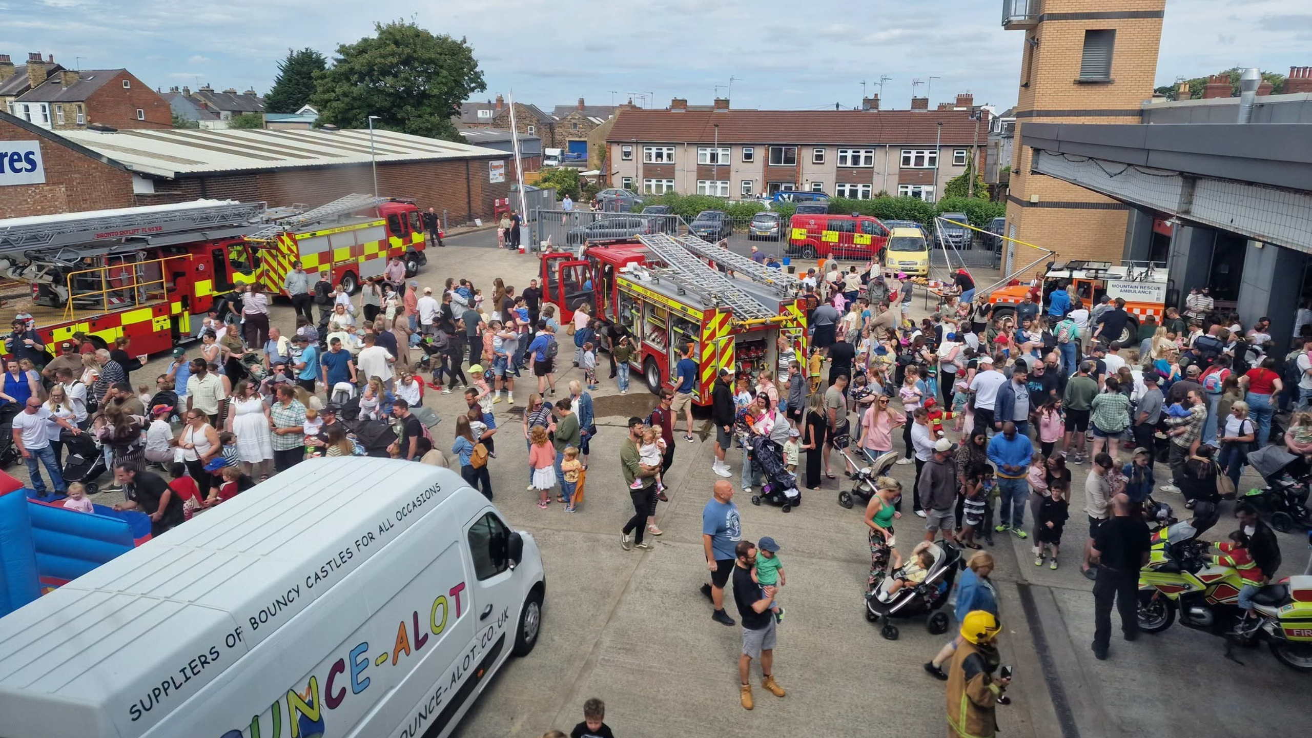 A large crowd, including families with children, gathers around fire trucks at an outdoor event. A white van labeled “Bounce-A-Lot” is in the foreground. Buildings and houses are visible in the background.