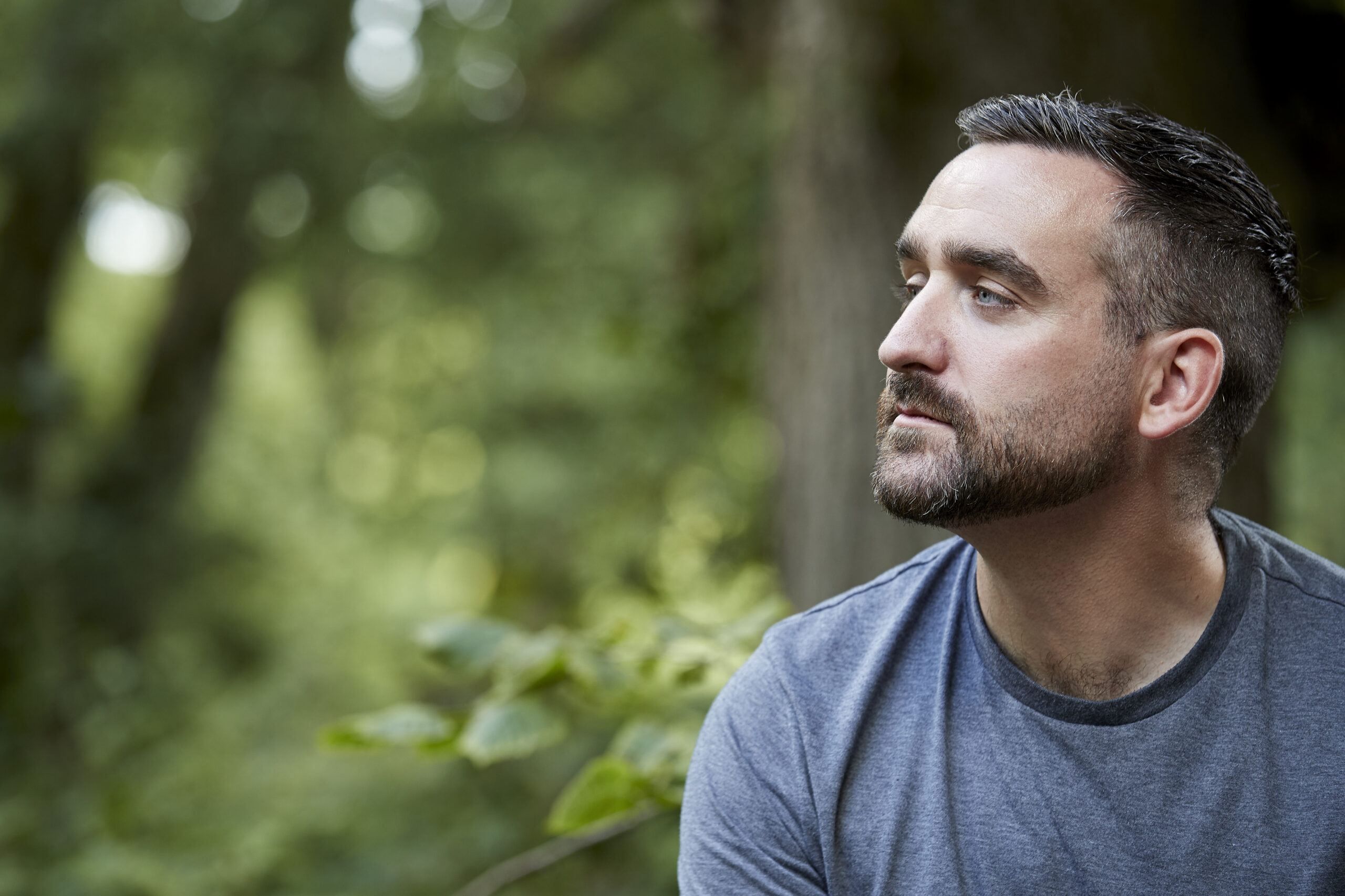 A man with short dark hair and a beard gazes thoughtfully to the right, sitting outdoors with green, blurred foliage in the background.