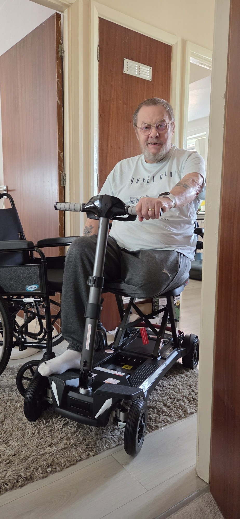 An older man with glasses smiles while sitting on a black mobility scooter indoors, next to a black wheelchair, in a hallway with wooden doors.