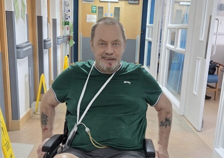A man in a wheelchair with a leg brace and red sock moves down a hospital hallway, wearing a green shirt and gray shorts. Medical equipment is attached to his body. Hallway signs and safety cones are visible.