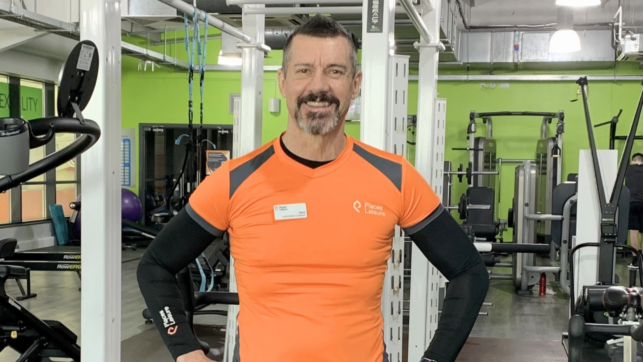 A man in an orange athletic shirt stands smiling with his hands on his hips in a gym, surrounded by various workout equipment and green walls.
