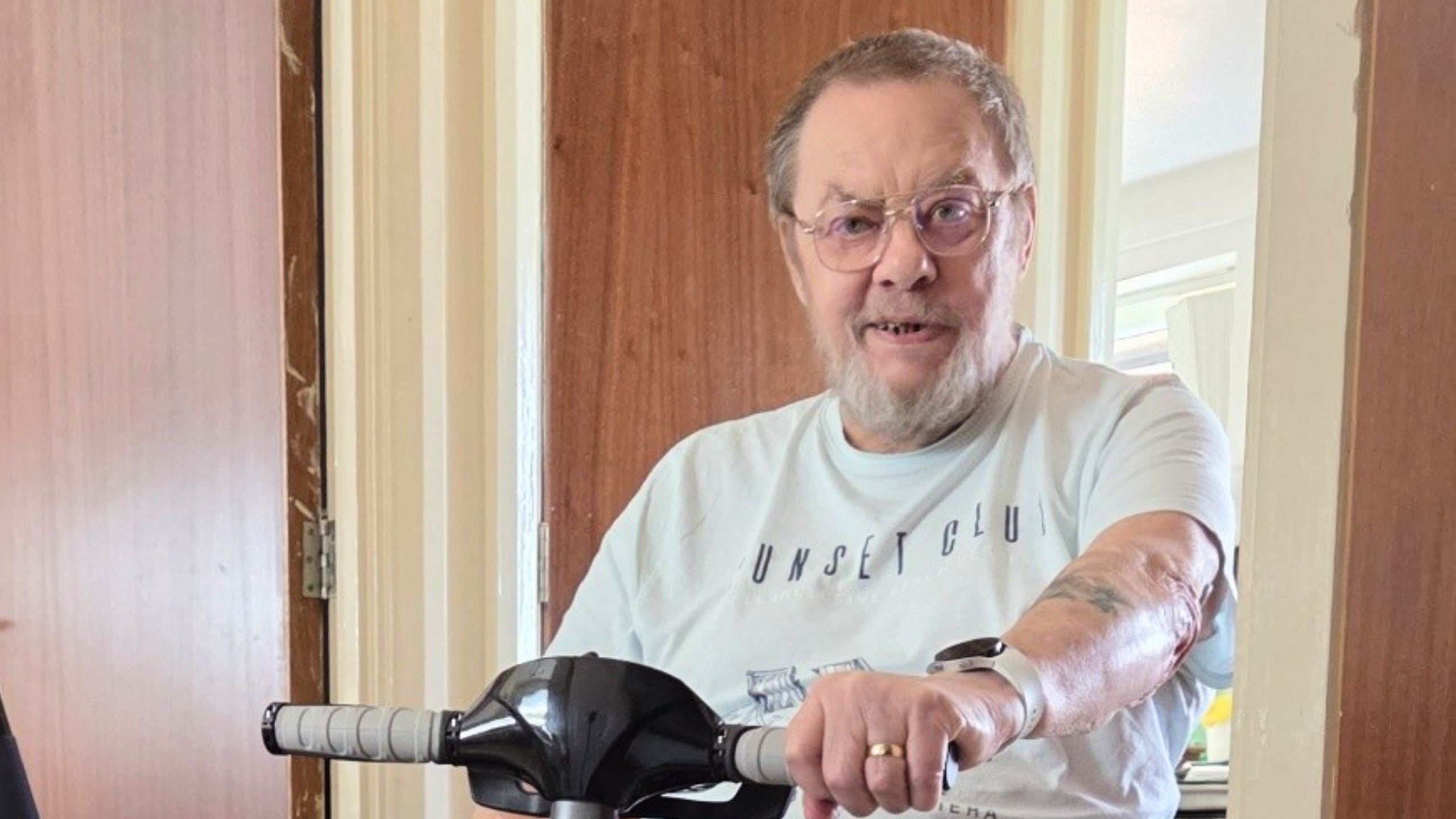 An older man with glasses and a beard sits indoors on a mobility scooter, smiling slightly. He is wearing a light-colored t-shirt and has visible tattoos on his right arm.