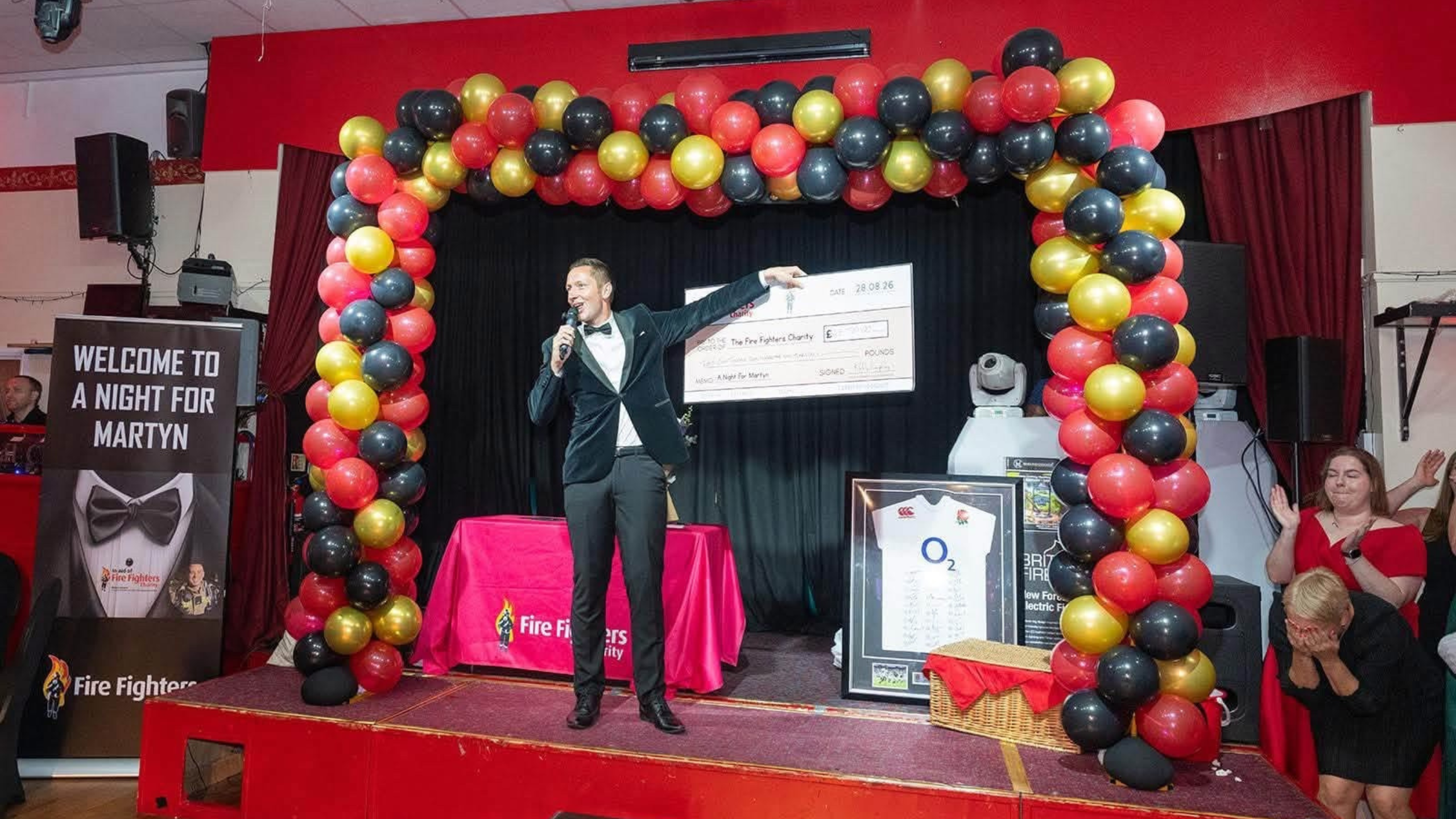 A man in a tuxedo holds a large check on stage under a red, black, and gold balloon arch at an event. A sign reads “Welcome to a Night for Martyn.” People applaud in the background.