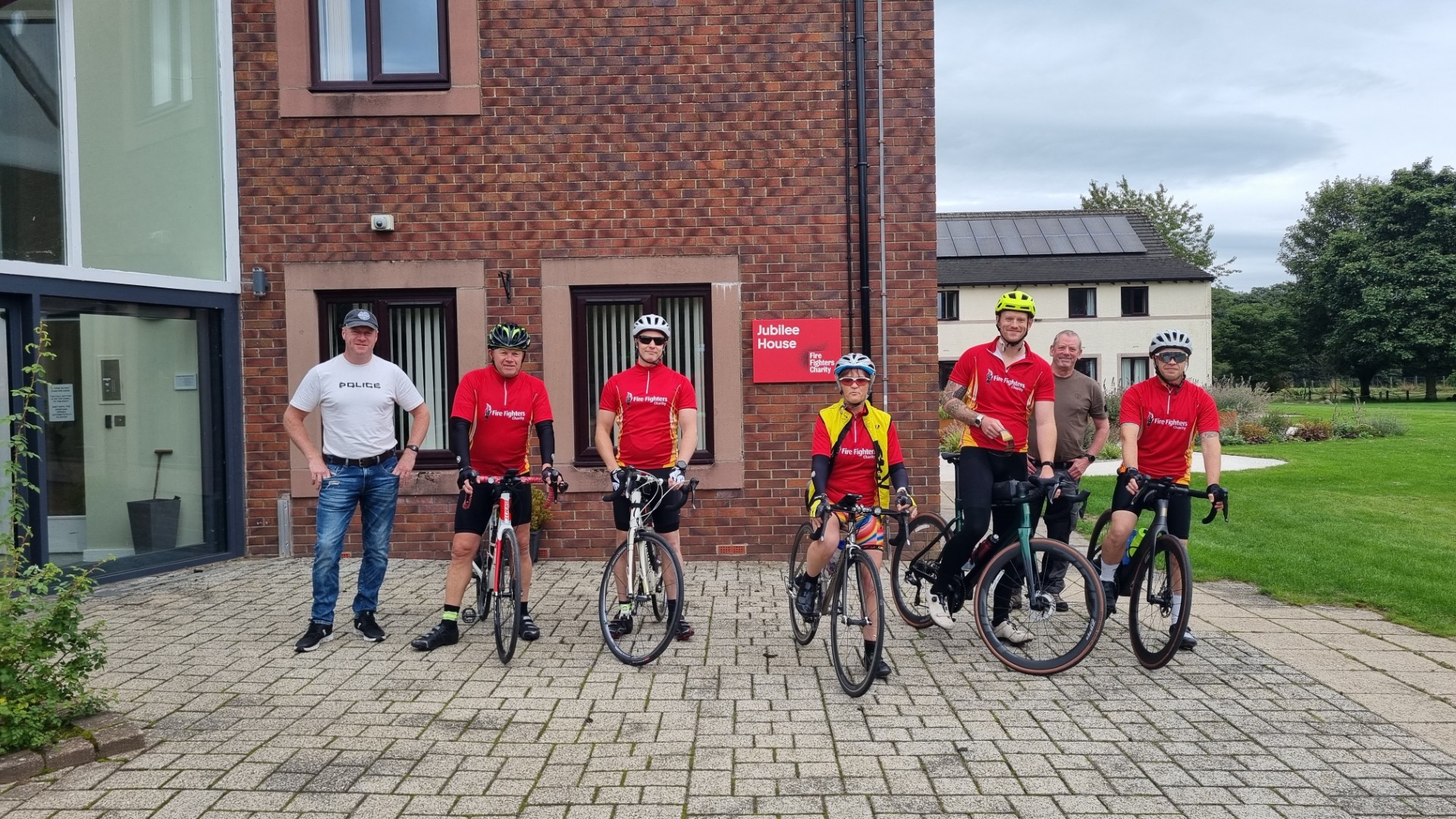 Six men pose outside a brick building; five wear helmets and cycling gear with bikes, while one in a white shirt and jeans stands on the left. A grassy area and another building are visible in the background.