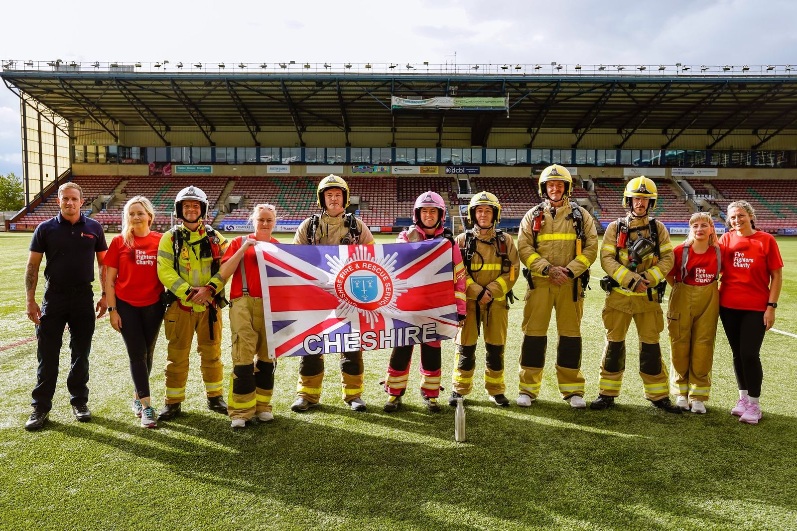 A group of people in firefighter gear and red shirts stand on a stadium field, holding a Cheshire Fire and Rescue Service flag with a Union Jack design.
