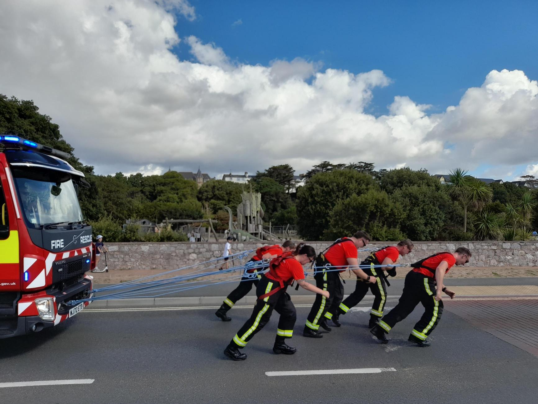 Five firefighters in uniform are pulling a fire truck with ropes during a public demonstration on a sunny day, with greenery and houses in the background.
