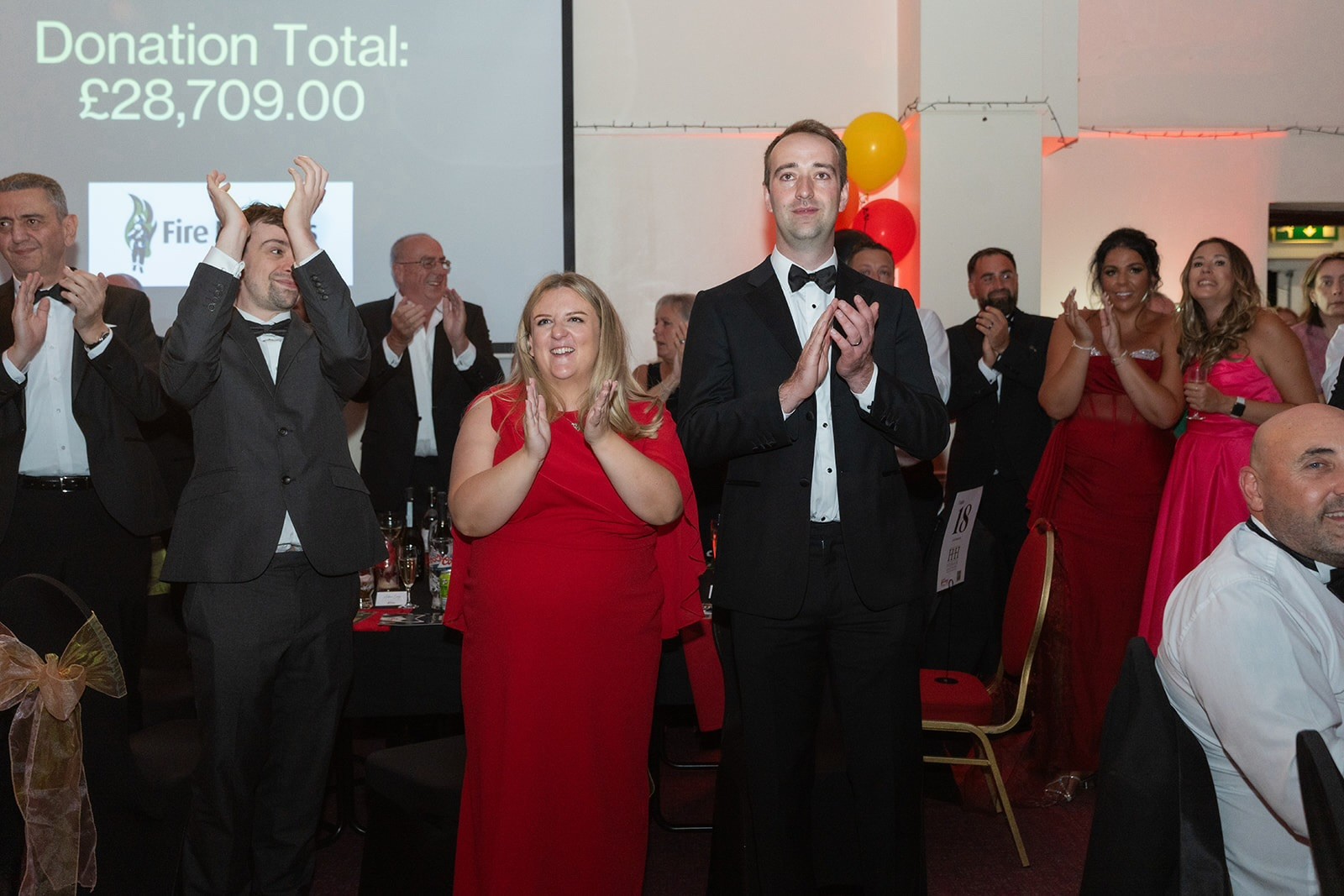 People in formal attire stand and applaud at an event, with a donation total of £28,709 shown on a screen in the background. Some are smiling and dressed in red dresses and black suits.