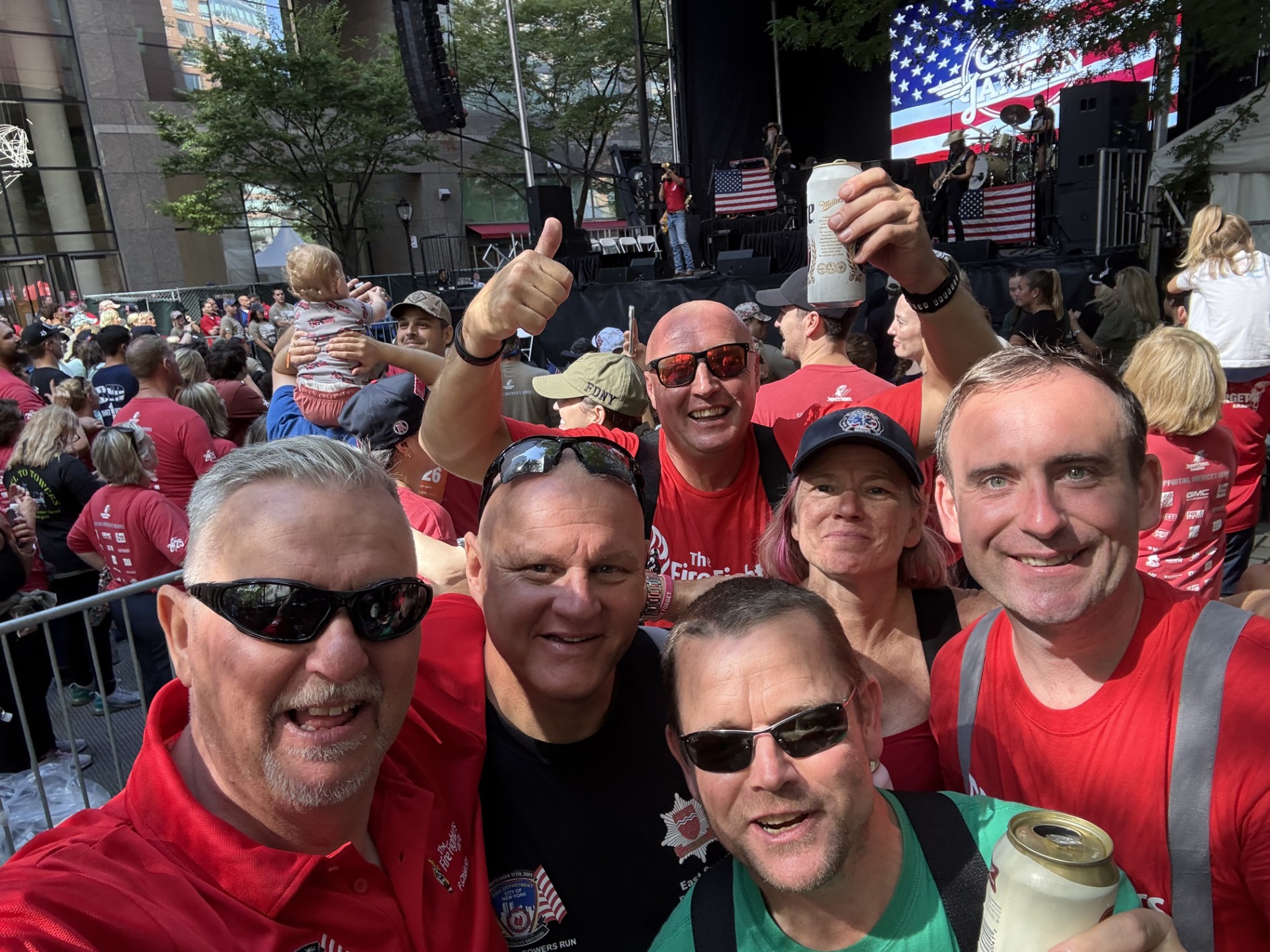 A group of six smiling people pose for a selfie at an outdoor event with a crowd, stage, and American flag in the background. Some hold drinks and wear red shirts. Trees and buildings surround the scene.