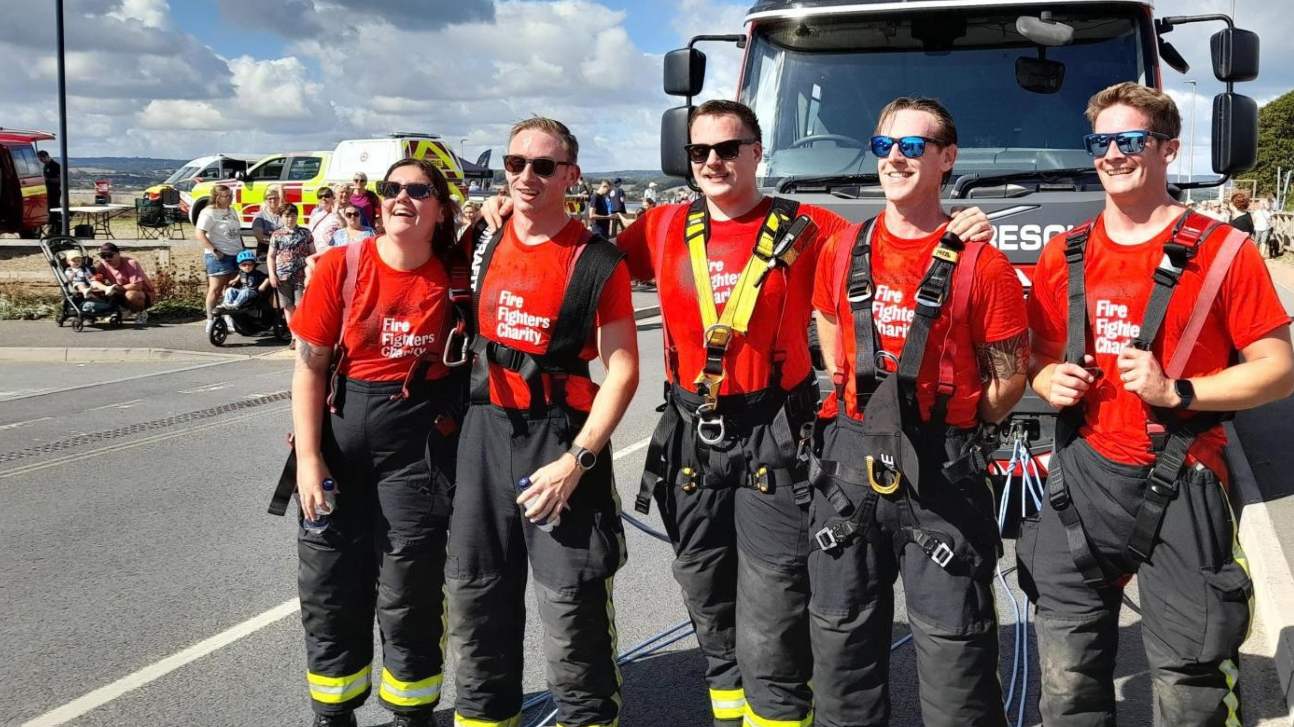 Five smiling people in red “Fire Fighters Charity” shirts and firefighter pants stand in front of a fire truck on a sunny day, with emergency vehicles and spectators in the background.