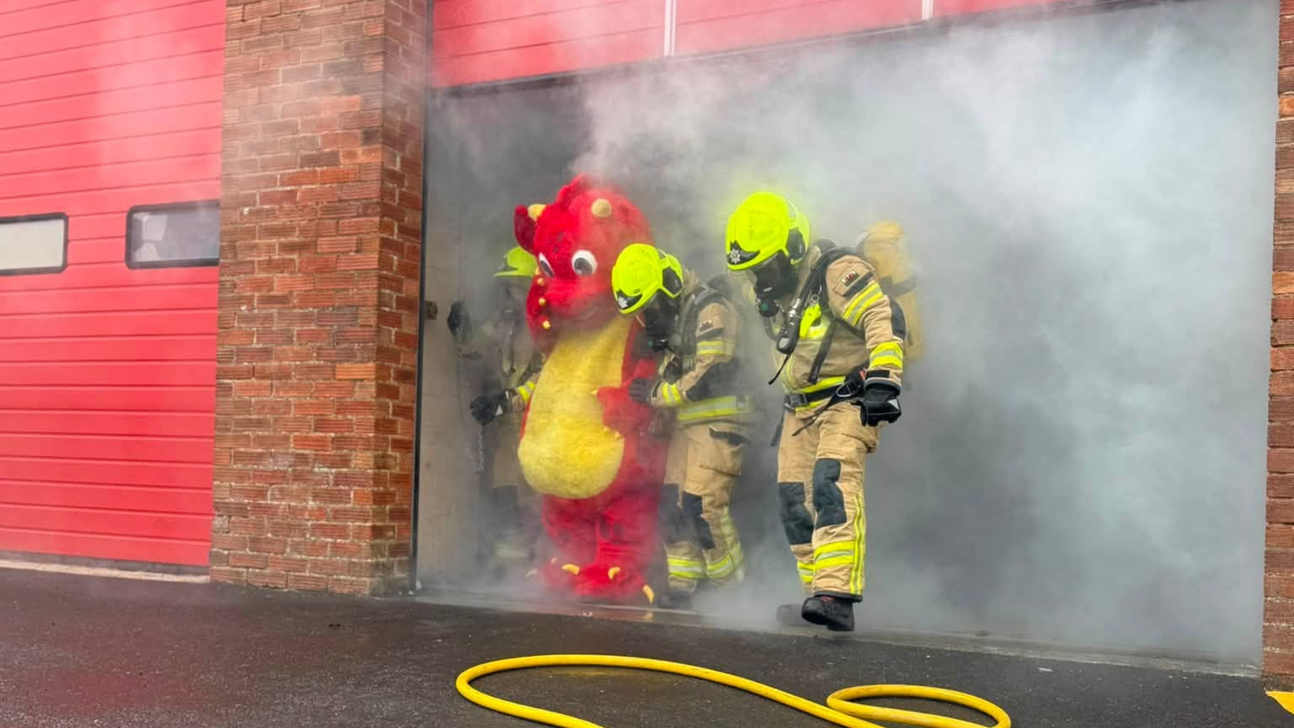 Three firefighters in gear guide someone in a red and yellow dragon costume out of a smoky building, next to a red garage door, with a yellow hose on the ground.