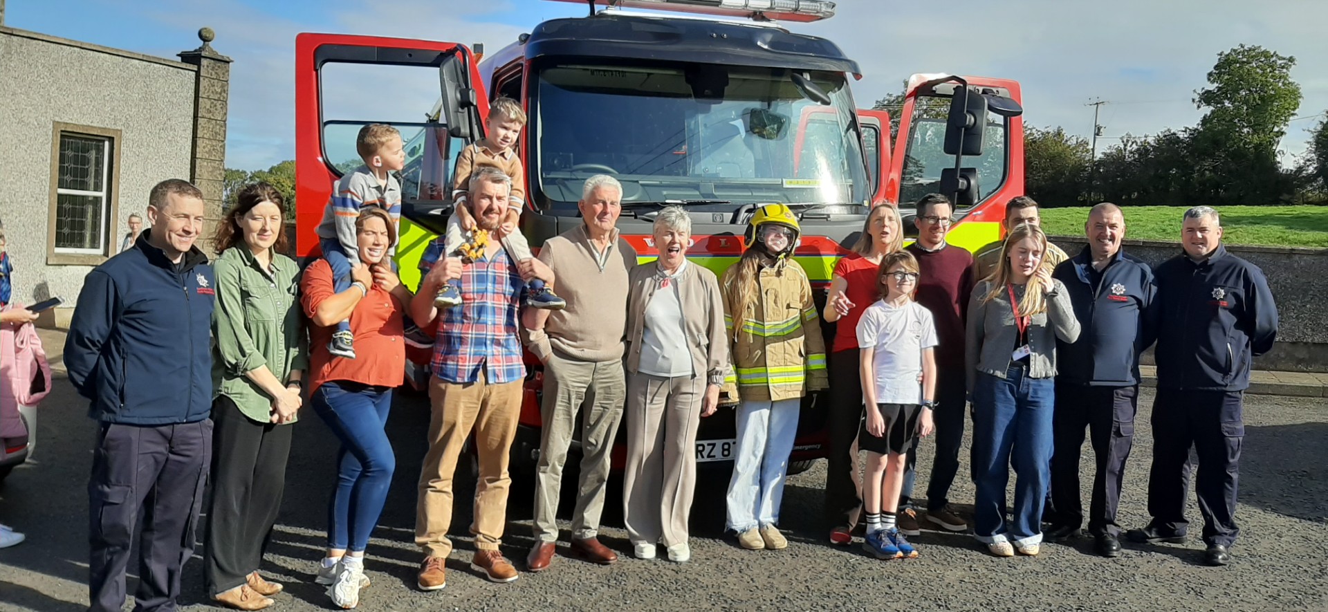 A group of adults and children posing for a photo in front of a red fire truck, with one child wearing a firefighter helmet.