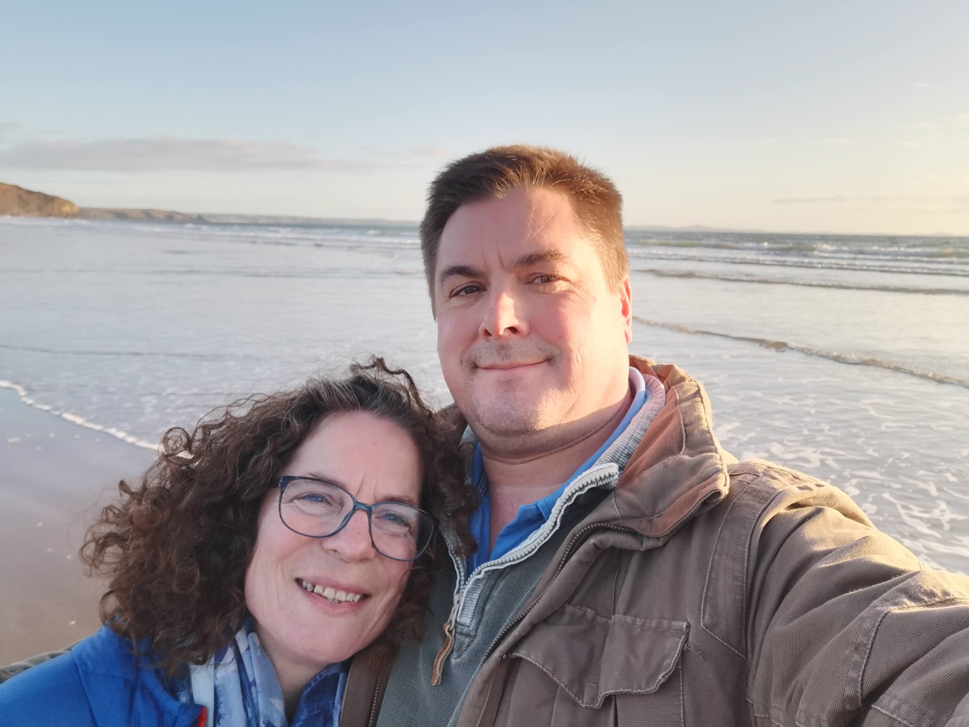 A man and a woman smile together while standing on a sandy beach at sunset. Waves are gently rolling in behind them, and the sky is mostly clear with soft light.