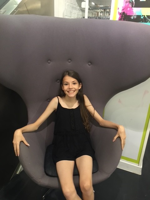 A smiling girl in a black sleeveless outfit sits in a large, modern, gray chair with high sides, her arms resting on the chair’s wide arms. Bright artwork and shelves are partially visible in the background.