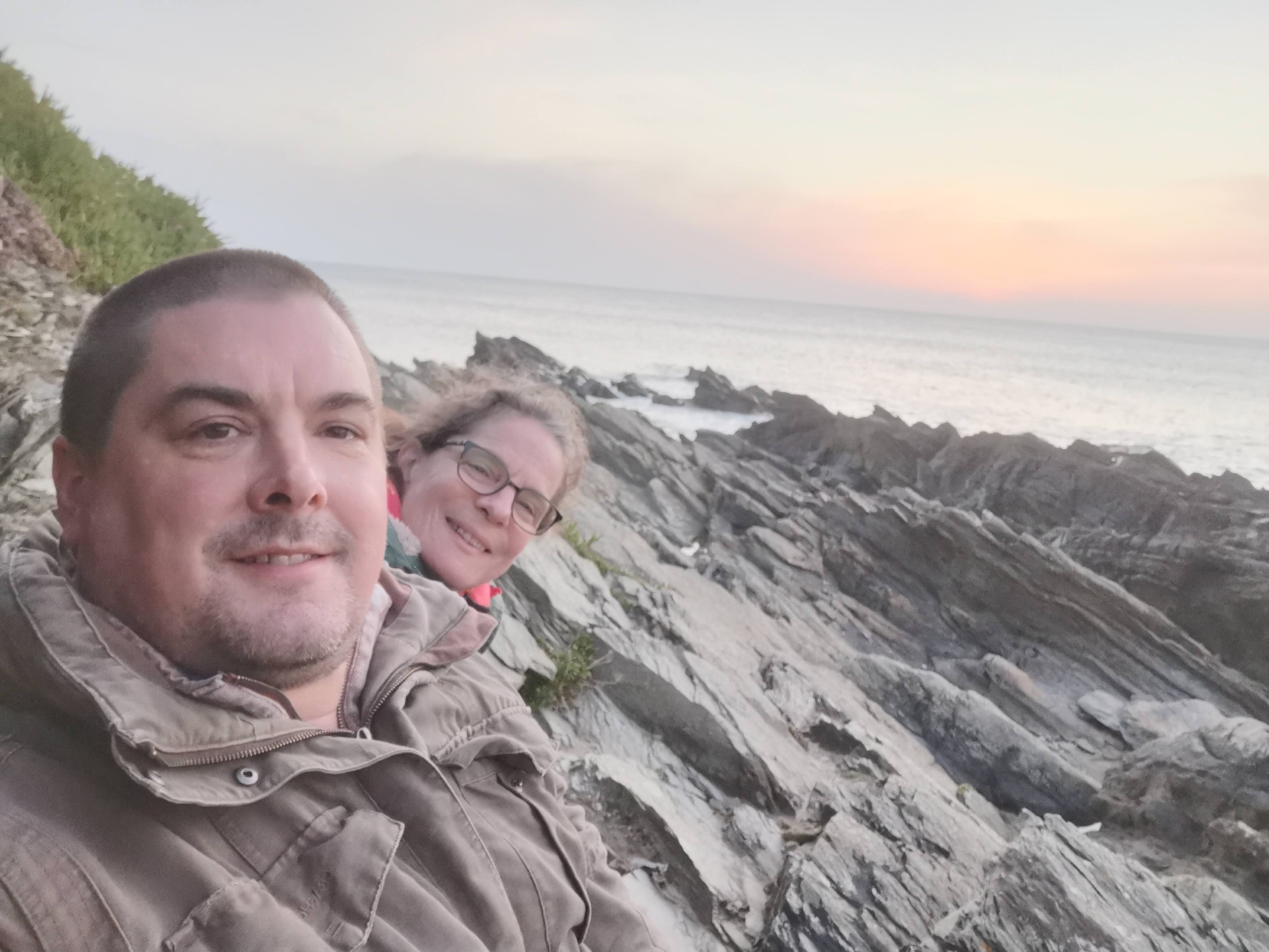 Two people in jackets smile at the camera while sitting on rocky cliffs by the sea at sunset, with gentle waves and a pastel sky in the background.