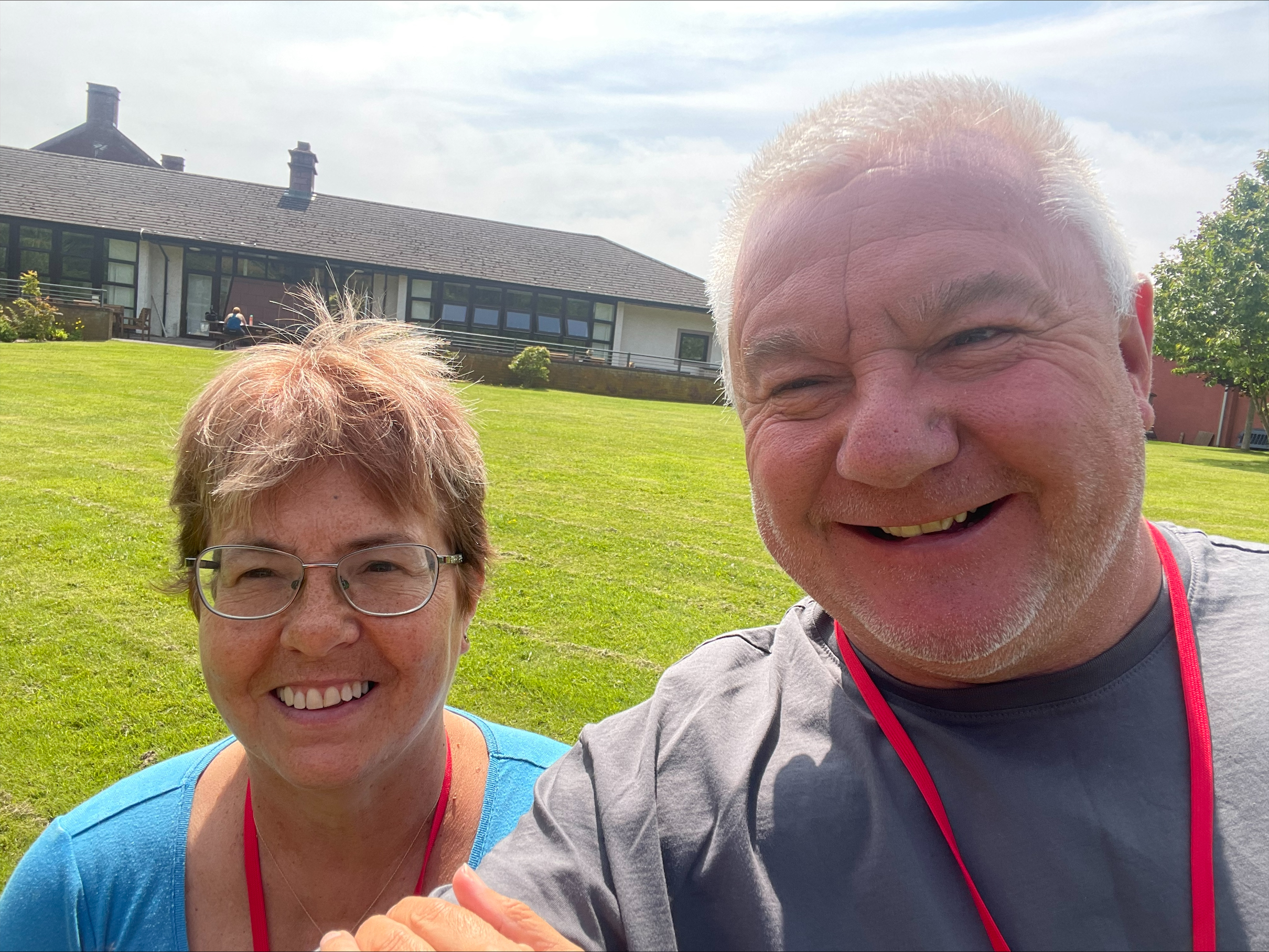 A smiling man and woman pose for a selfie on a sunny lawn in front of a house.