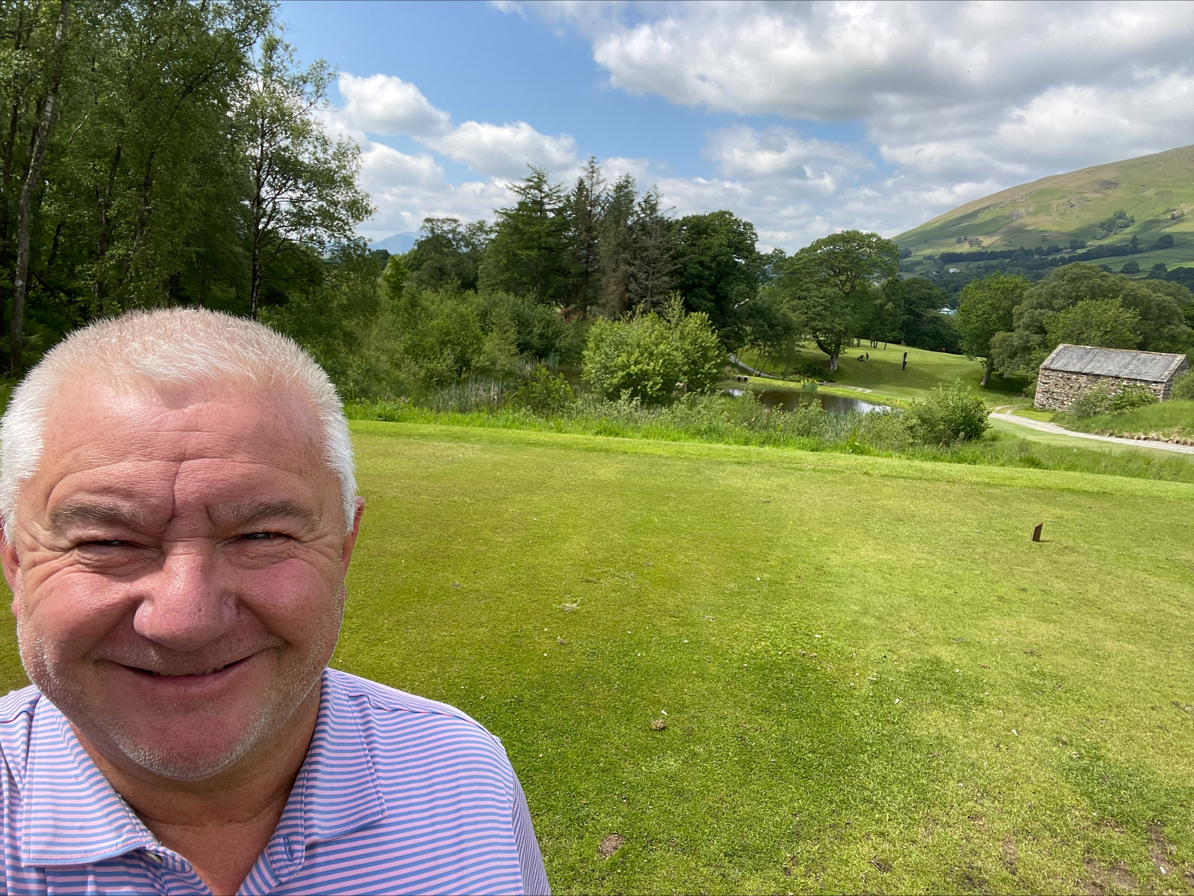 A smiling man takes a selfie on a green golf course, with a pond and hills in the background.