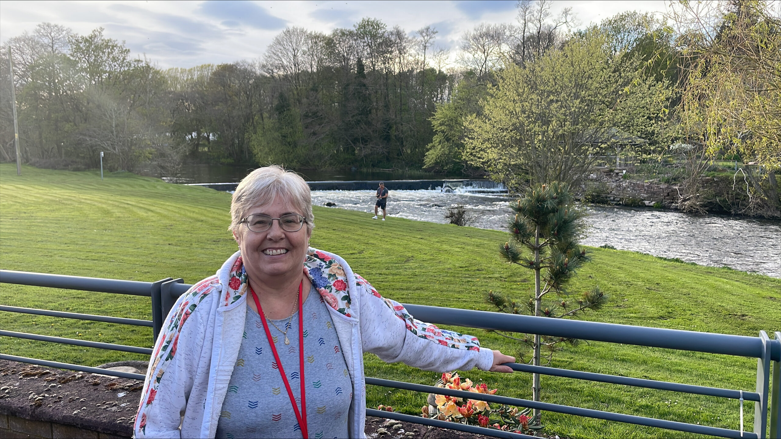 A smiling woman stands in front of a river with a small waterfall, with a grassy park area in the background.