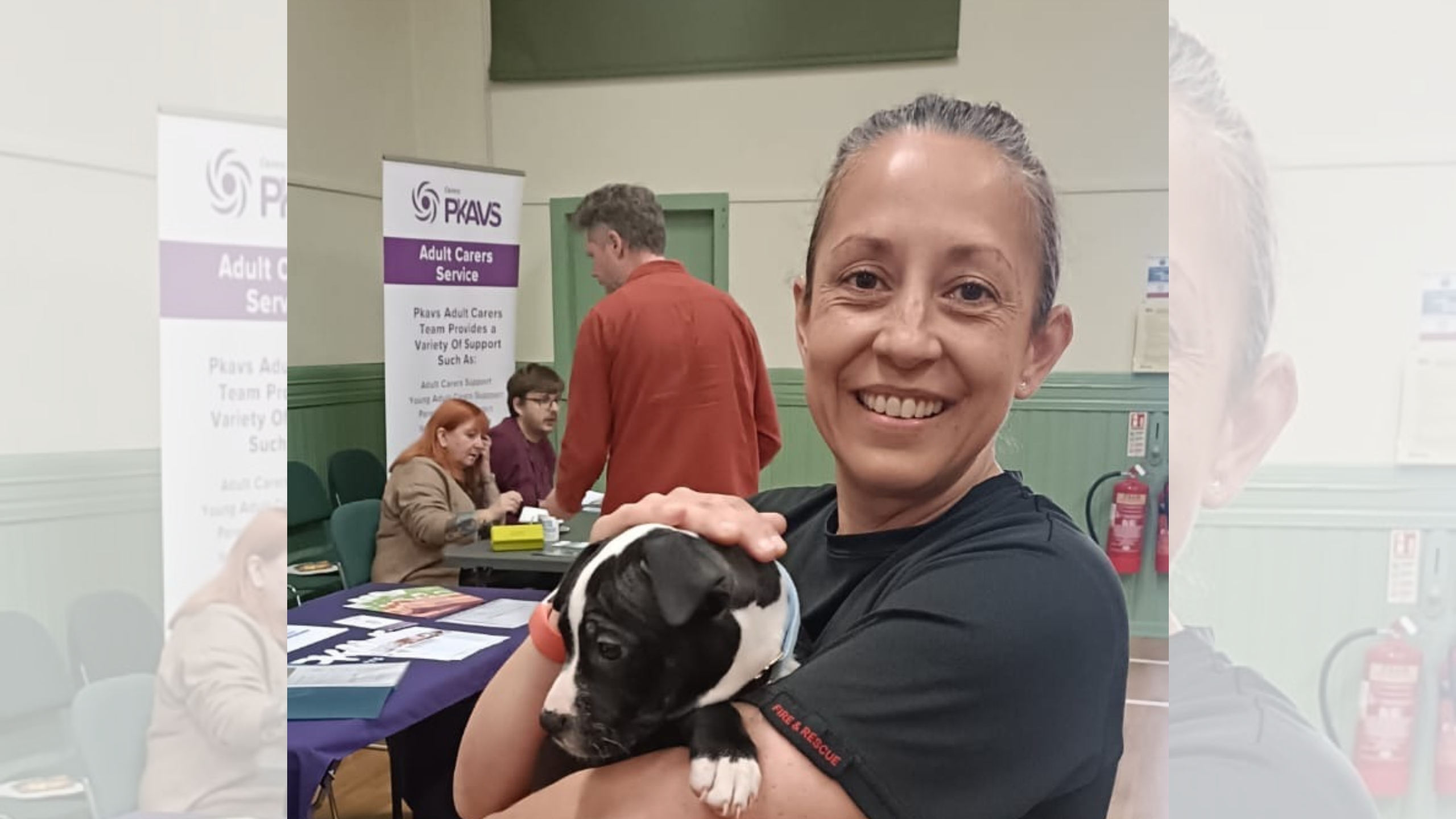 A woman in a black uniform shirt holds a small black and white puppy while standing in front of a PKAVS Adult Carers Service banner.
