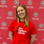 A smiling woman wearing a red Fire Fighters Charity t-shirt stands in front of a red backdrop featuring the Northern Ireland Fire & Rescue Service logo as part of our regional fundraisers team.