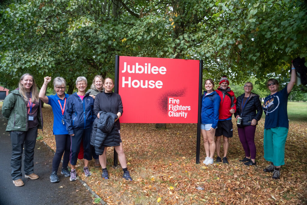 A group of eight people standing in front of a red sign that reads “Jubilee House, Fire Fighters Charity.”