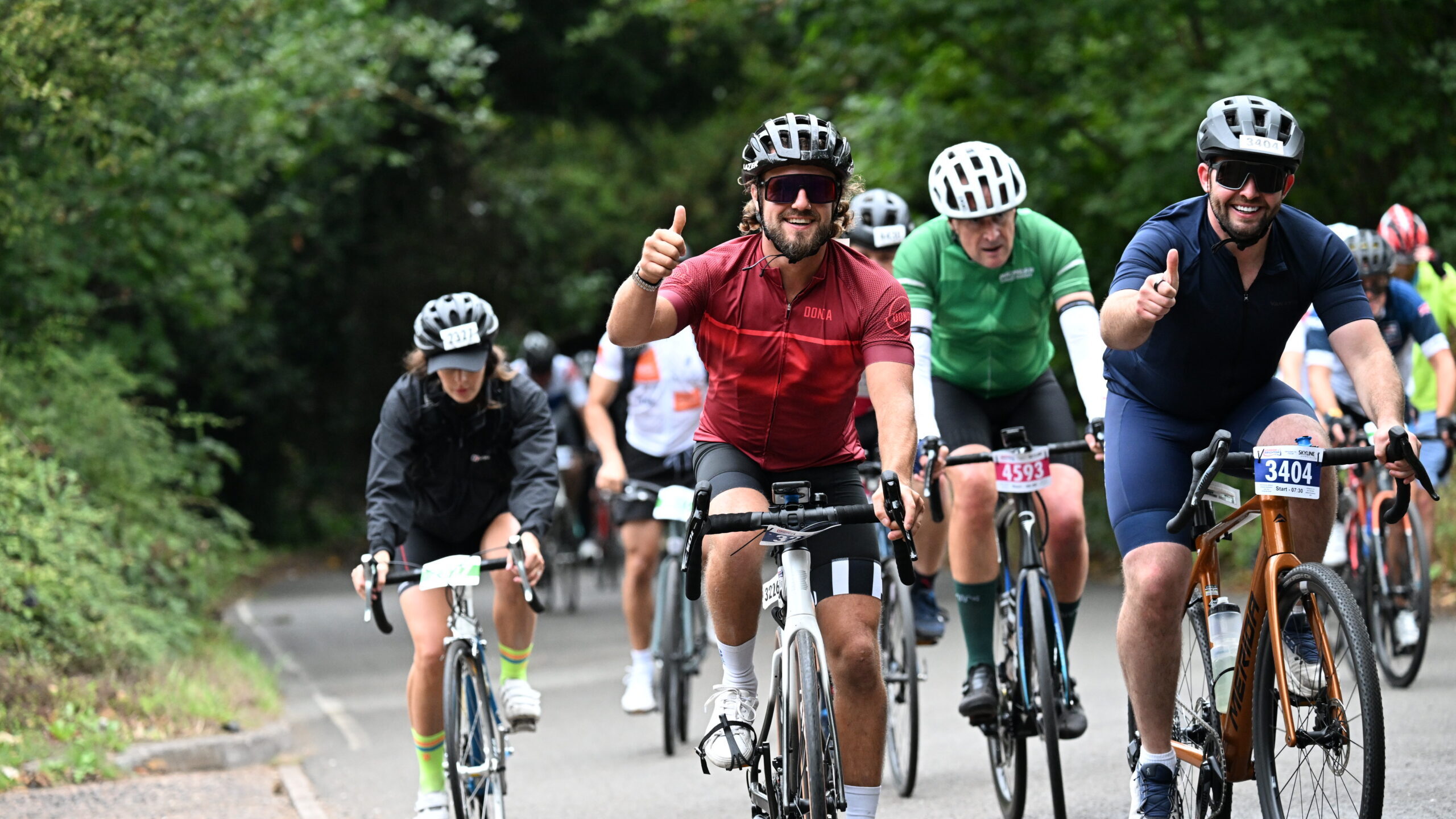 Cyclists riding on a road, with two men in the foreground giving a thumbs up.