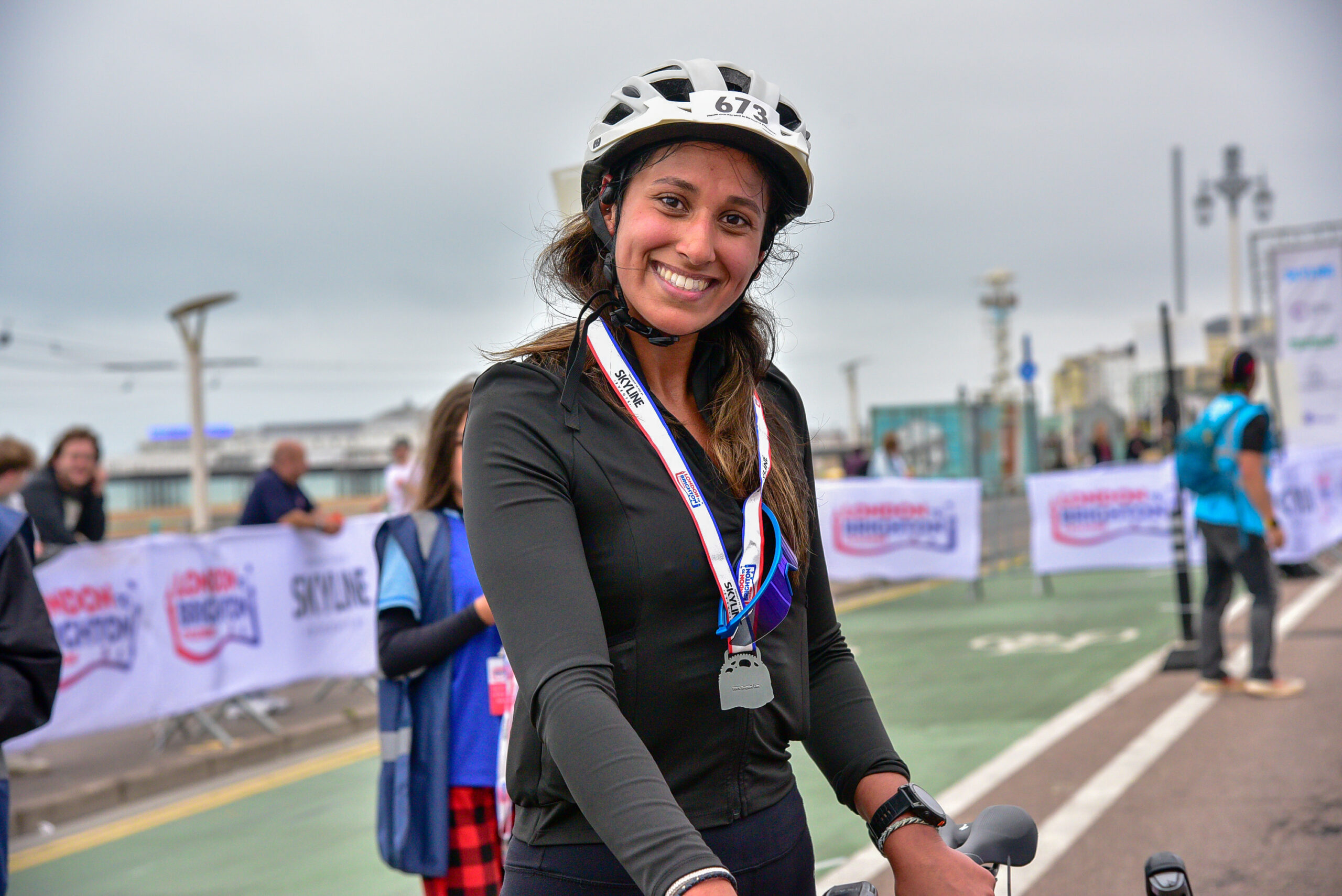 A smiling cyclist wearing a helmet and a race medal around her neck stands on a path lined with event banners. 