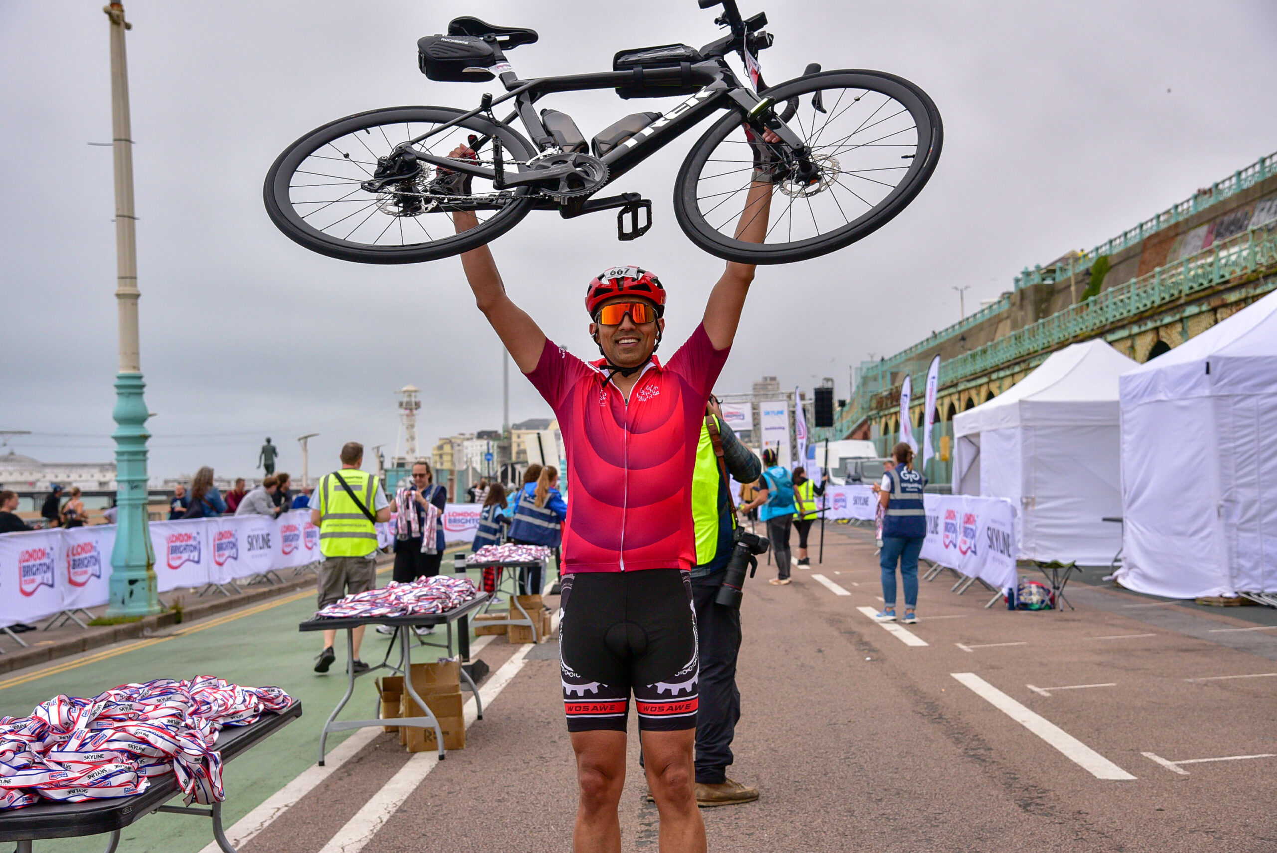 A cyclist in a red jersey and helmet holds their bike above their head in celebration on a beachfront path. 
