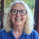 A smiling woman with shoulder-length white hair wearing black-rimmed glasses and a blue collared shirt, part of the fire fighters charity fundraising team.