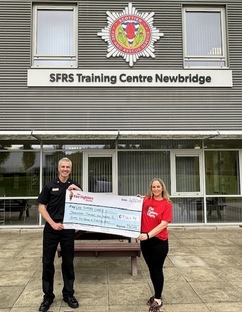 A man in a Scottish Fire and Rescue Service uniform and a woman in a red Fire Fighters Charity t-shirt hold a large donation check outside the SFRS Training Centre Newbridge.