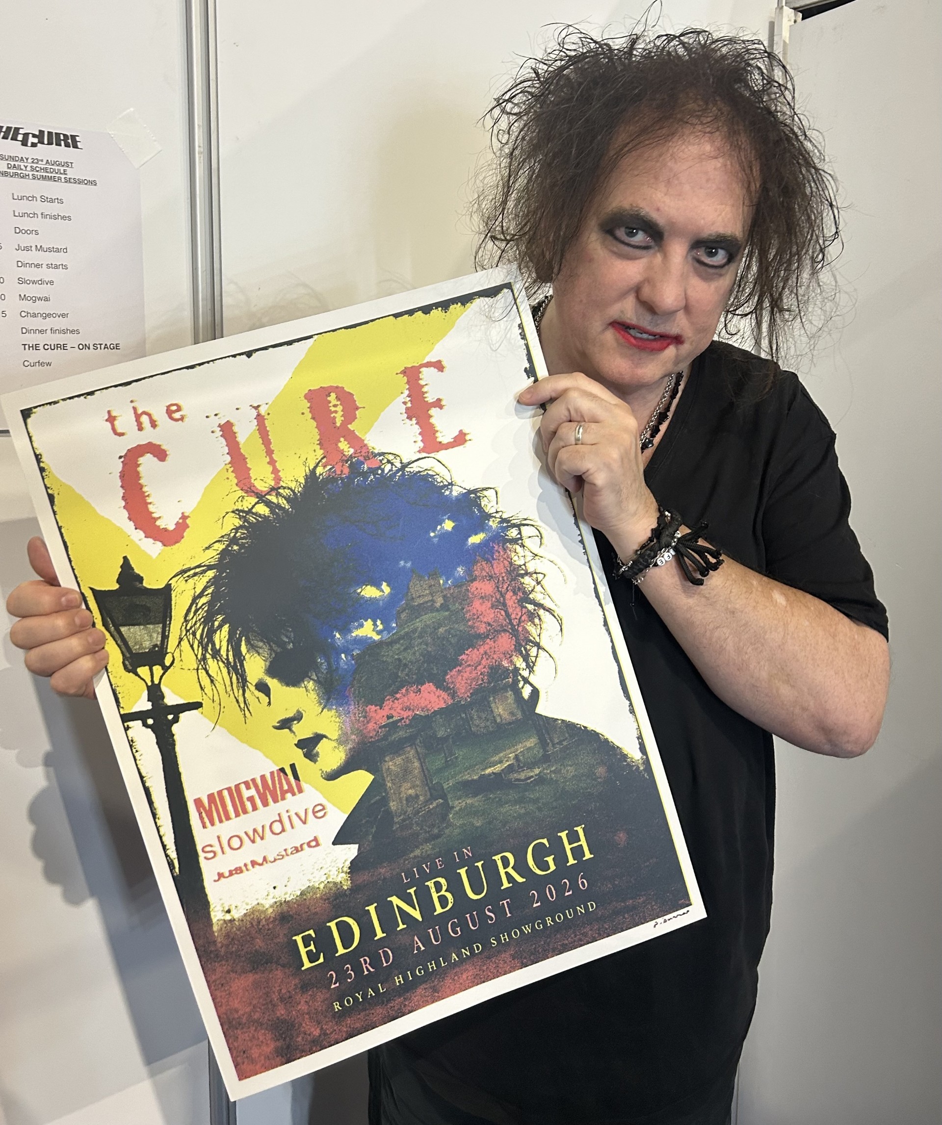 Robert Smith holding a concert poster for The Cure, live in Edinburgh on August 23, 2026, with opening acts Mogwai, Slowdive, and Just Mustard.