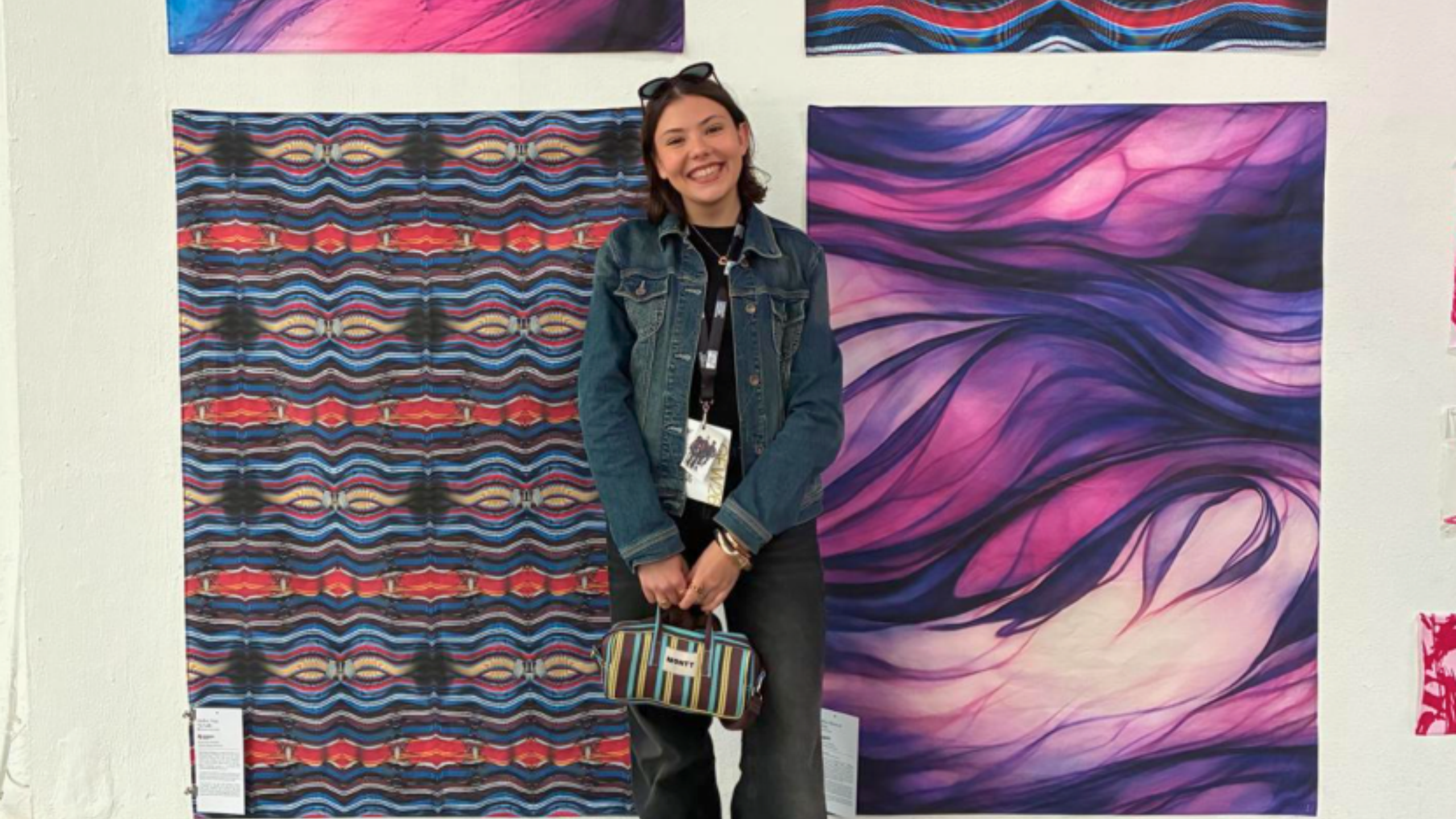 A woman in a denim jacket smiles and stands in front of two colorful abstract artworks—one with wavy purple patterns and one with red, blue, and white zigzags—displayed on a white wall. She holds a striped bag.