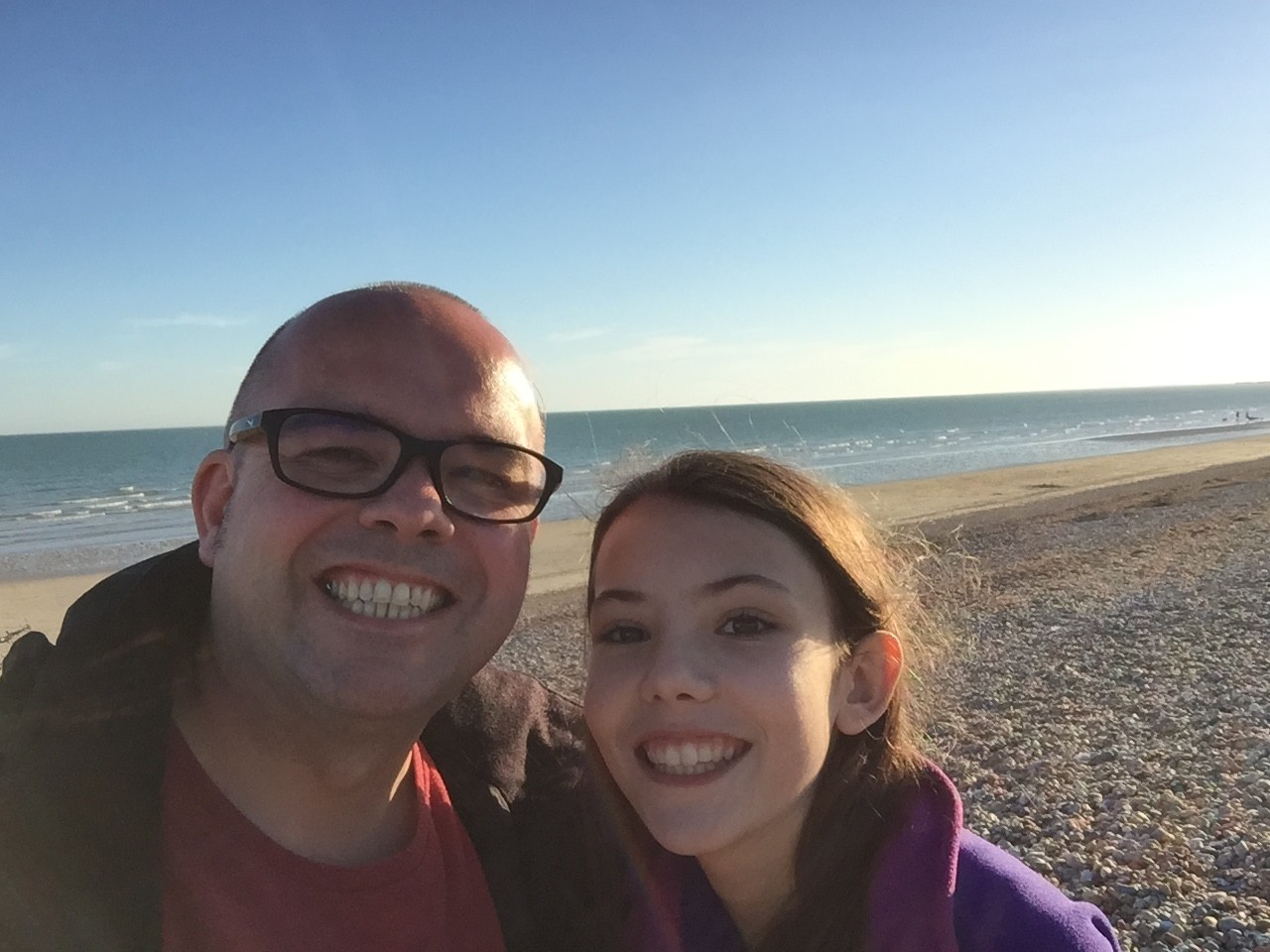 A man and a young girl smile at the camera while standing on a rocky beach with the ocean and blue sky in the background.