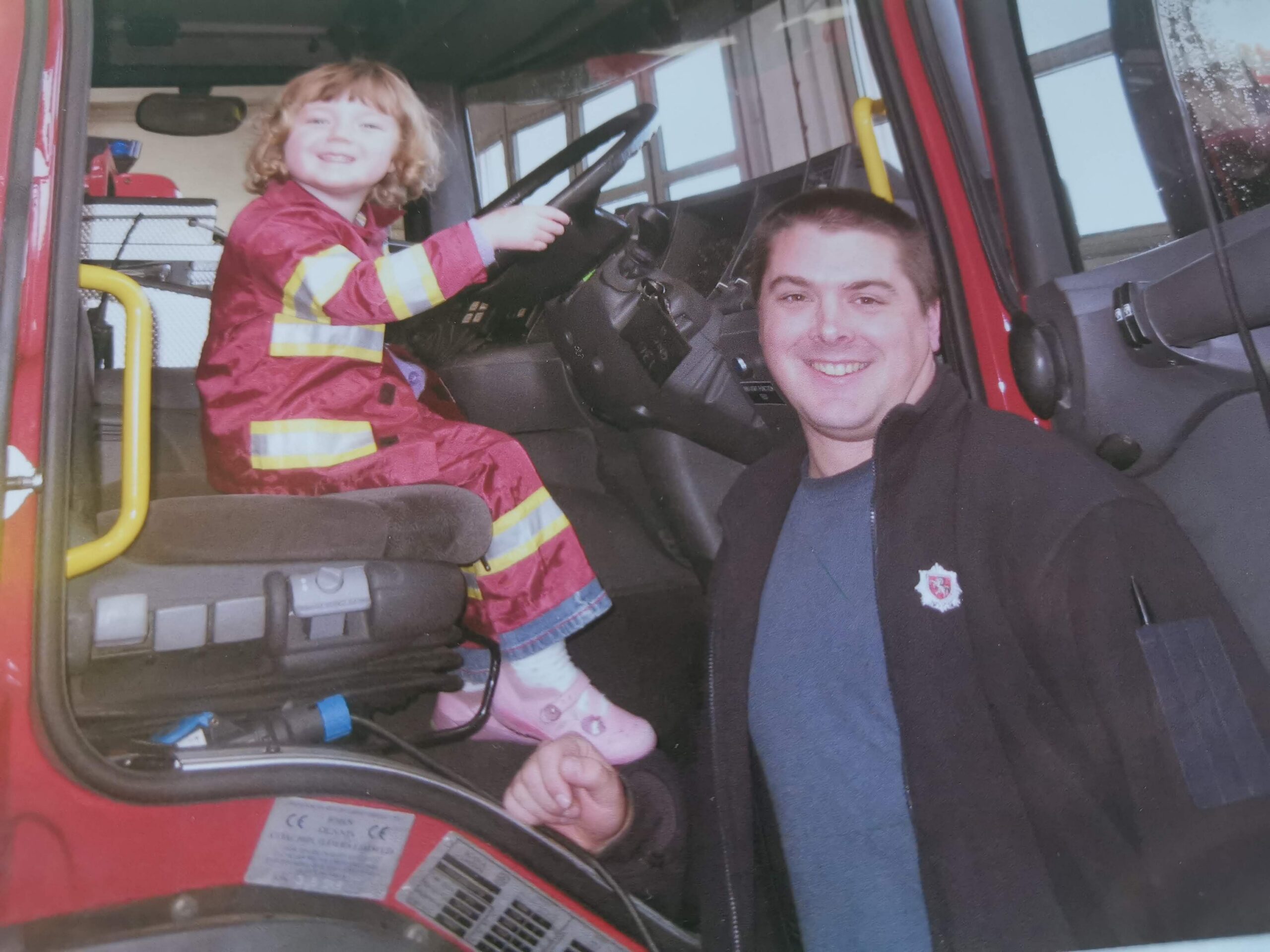 A smiling young child in a firefighter outfit sits in a fire truck’s driver seat, holding the steering wheel. An adult in a dark jacket with a fire department logo stands next to the door, smiling.