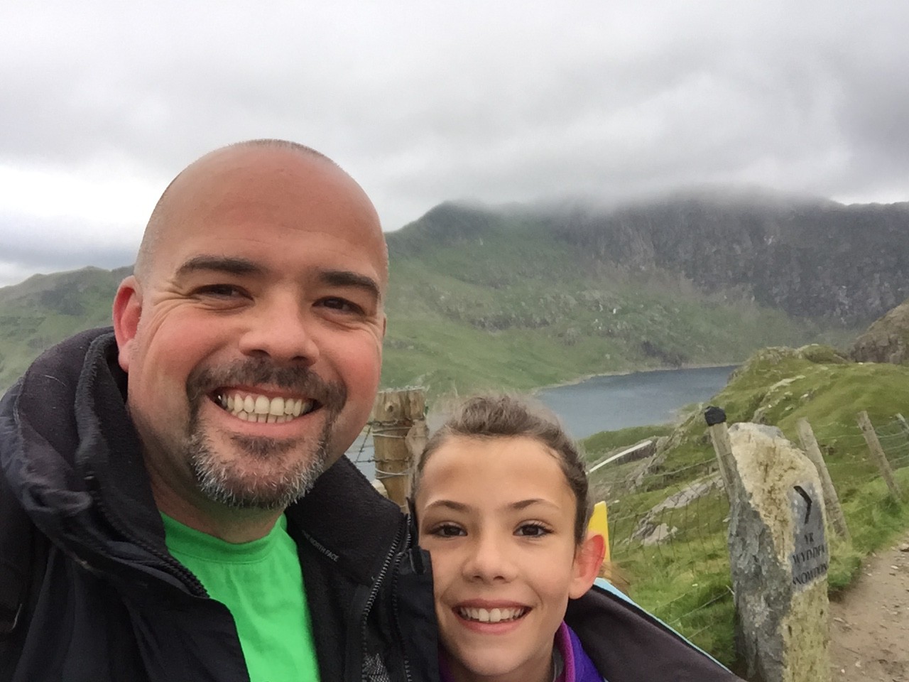 A man and a child smiling for a selfie outdoors with green hills, rocky terrain, and a lake in the background under a cloudy sky.