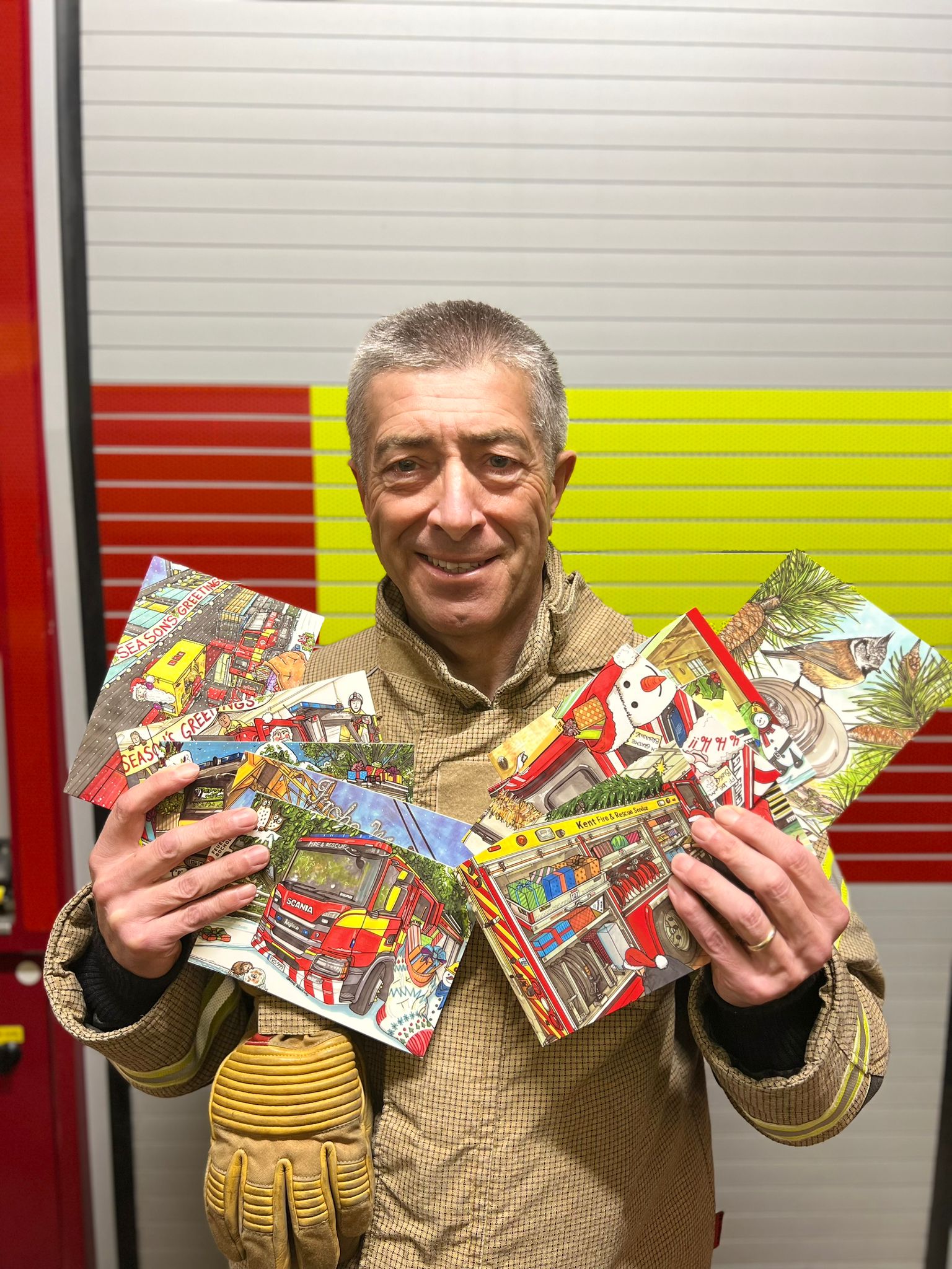 A smiling firefighter in uniform holds up a collection of illustrated holiday cards featuring fire trucks and winter scenes.
