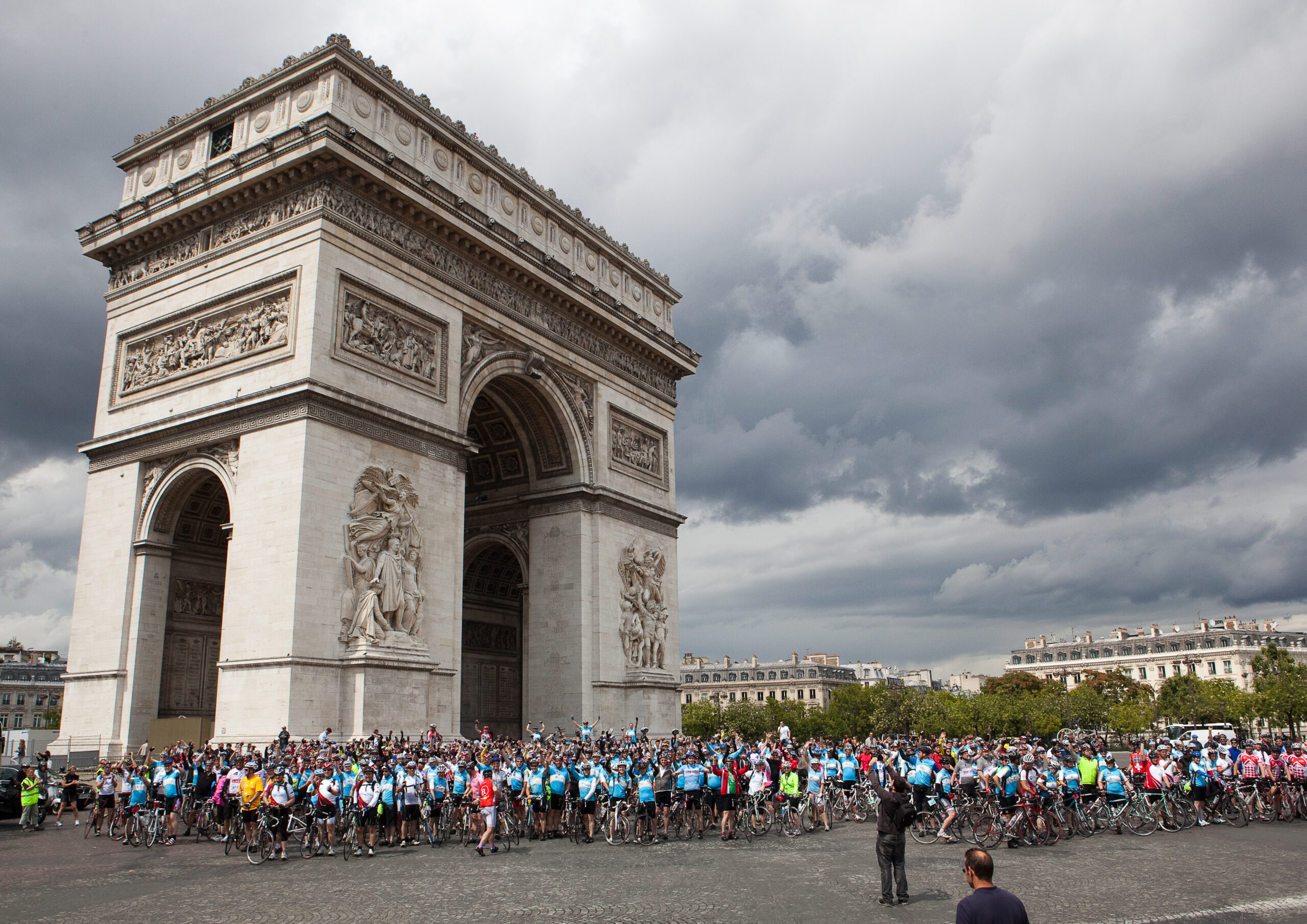 A large group of cyclists in blue jerseys gather in front of the Arc de Triomphe in Paris under a cloudy sky.