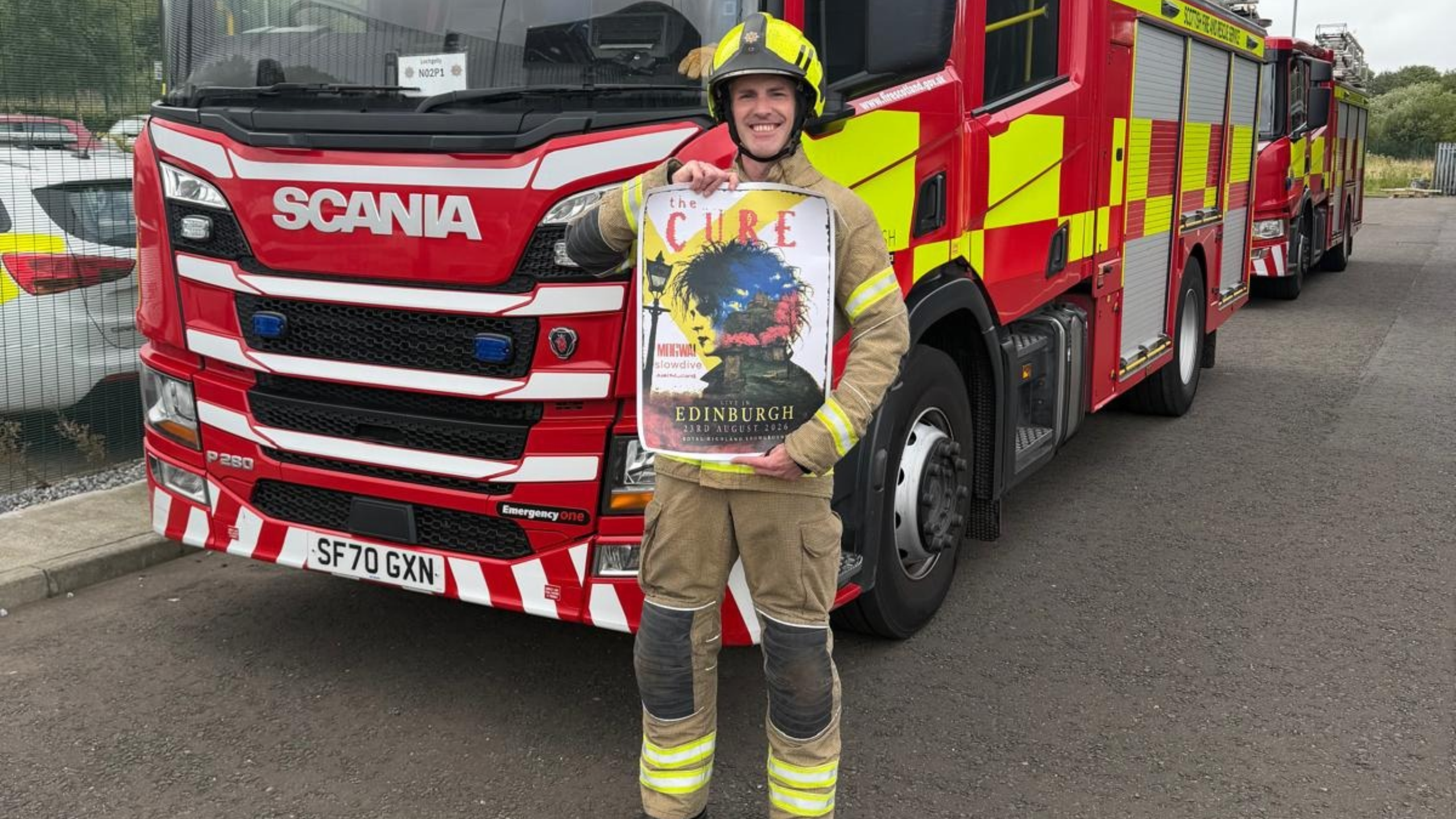 A firefighter in uniform holds a concert poster for The Cure in Edinburgh in front of a red Scania fire engine.